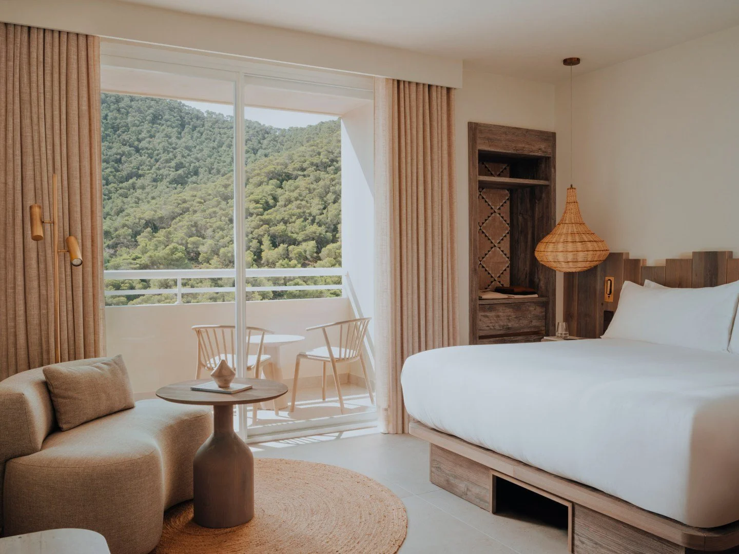 Modern hotel room with a large bed, cozy sofa, wicker pendant lamp, and sliding glass doors leading to a balcony with forested view.