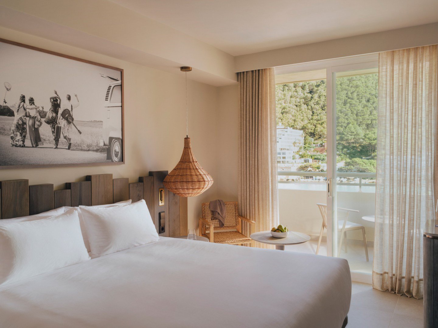Modern hotel room with a large bed, wicker pendant lamp, wall art, and a balcony with a view of trees. Includes a small table with a wicker chair and bowl of fruit.