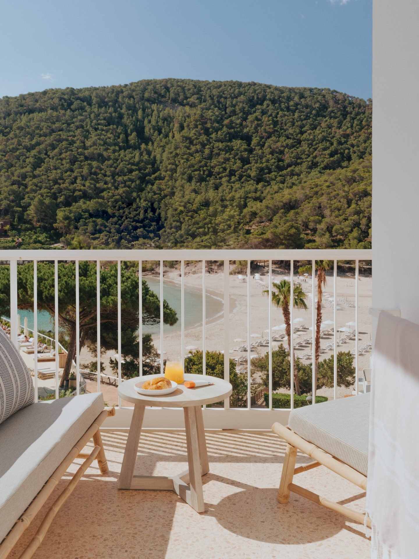 Scenic view from a balcony with two lounge chairs, a side table with breakfast items, overlooking a beach and wooded hillside.