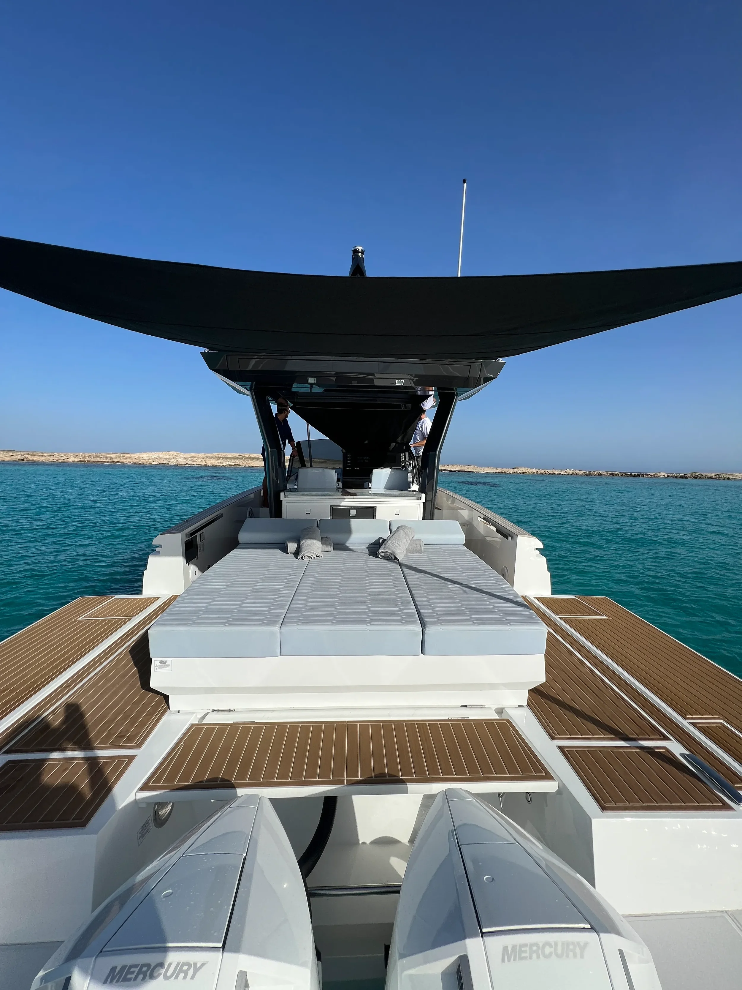 View from the stern sunbed of the Fiart 35 Martone W with the black sunshade extended over the deck while anchored in clear turquoise water.