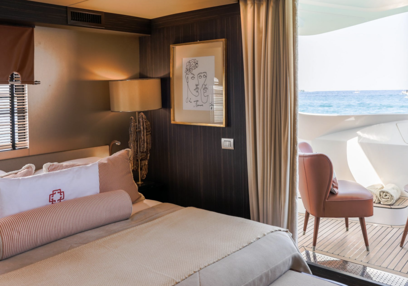 Luxury yacht cabin interior with ocean view, elegant bed, modern art on wall, stylish chair outside on deck.