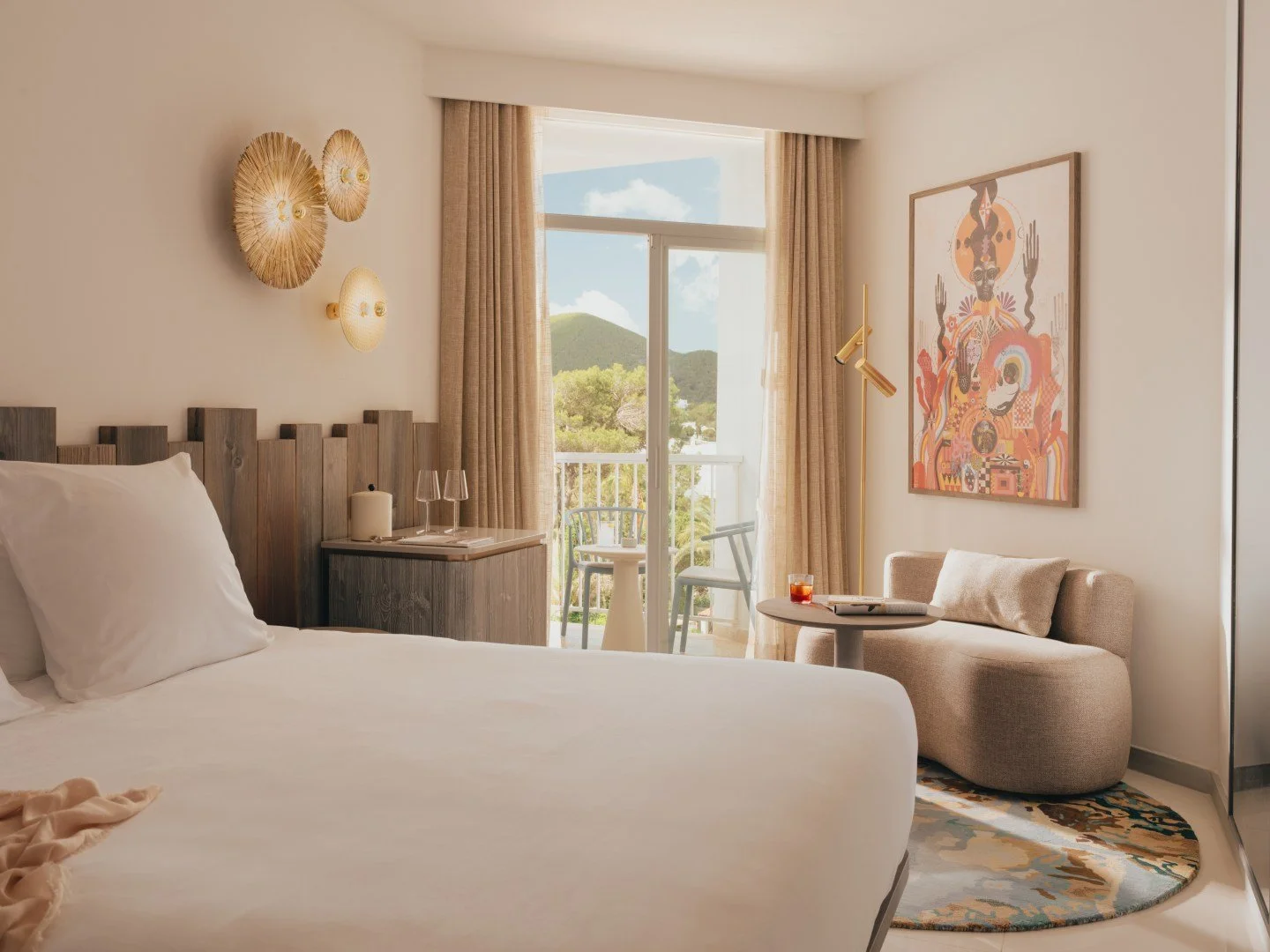 Modern hotel room with a cozy bed, wooden headboard, wall art, circular wall lamps, a small round coffee table, beige sofa, colorful rug, and a balcony with outdoor seating.