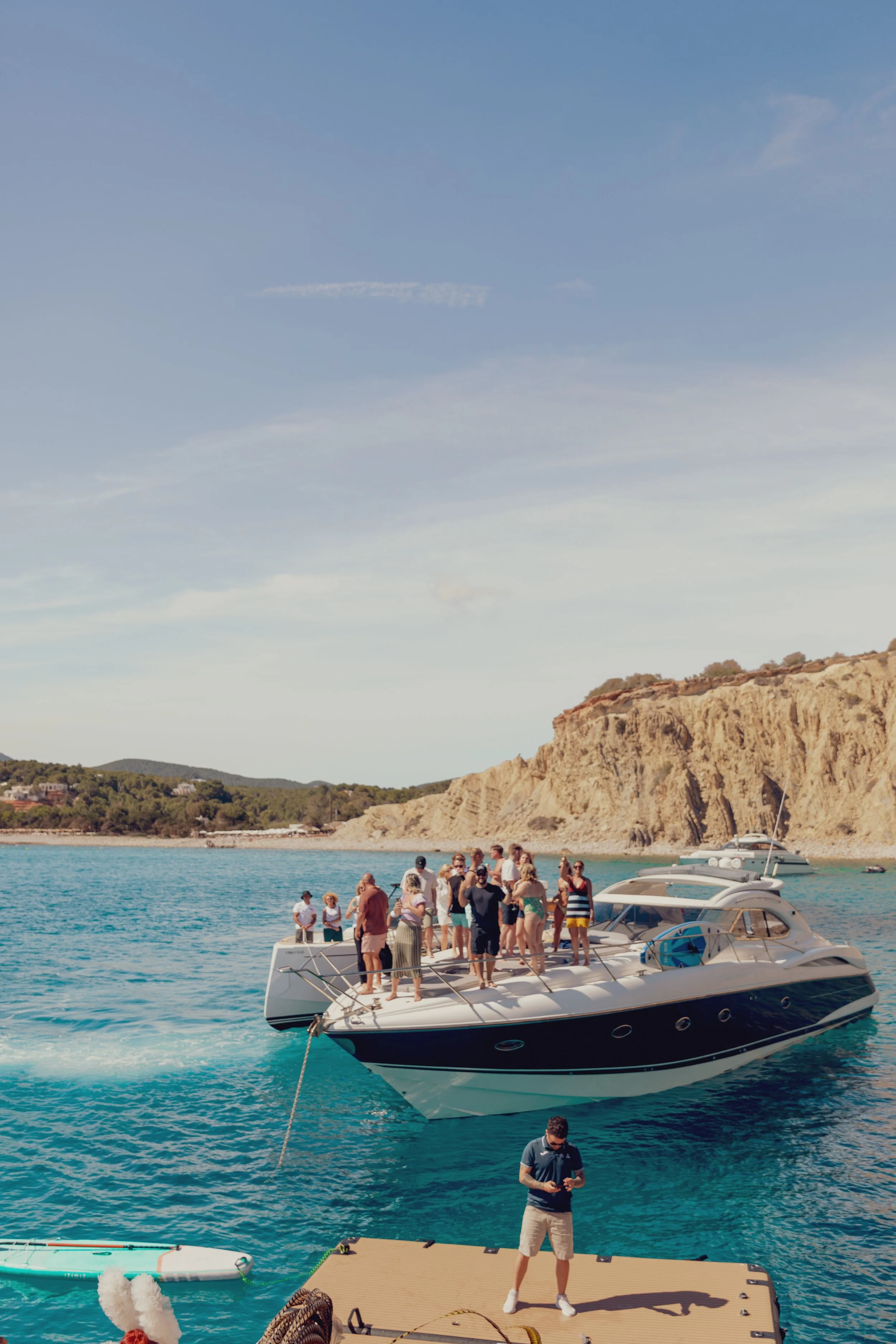 A group of people enjoying a boat party on a yacht in a turquoise sea near rocky cliffs under a clear sky.