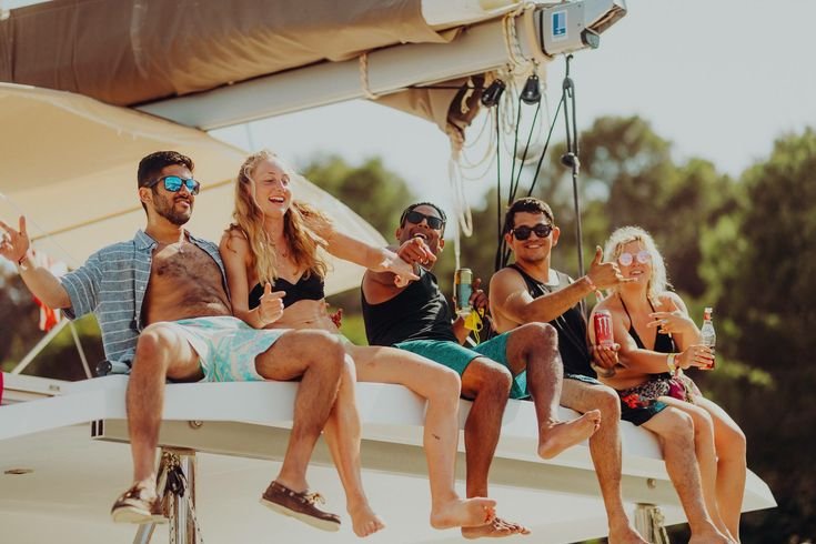 Group of five friends enjoying a boat ride on a sunny day, sitting on the edge of a sailboat, smiling, making peace signs, and holding drinks.