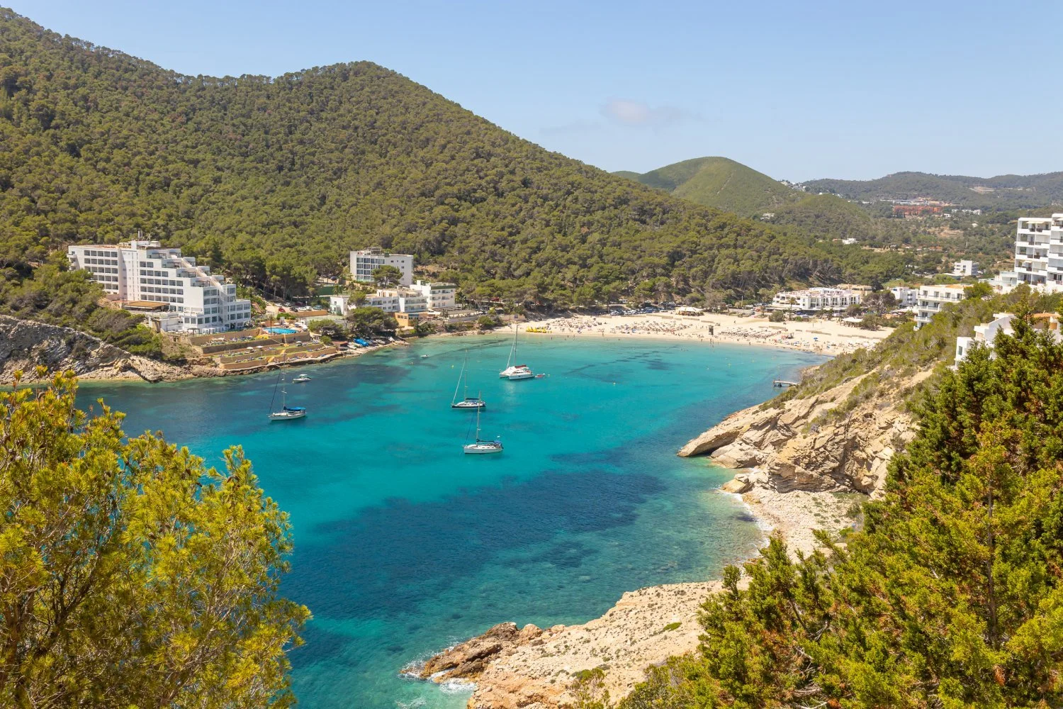 Scenic view of Cala Vadella beach, Ibiza, with turquoise waters, sailing boats, surrounding green hills, and residential buildings.