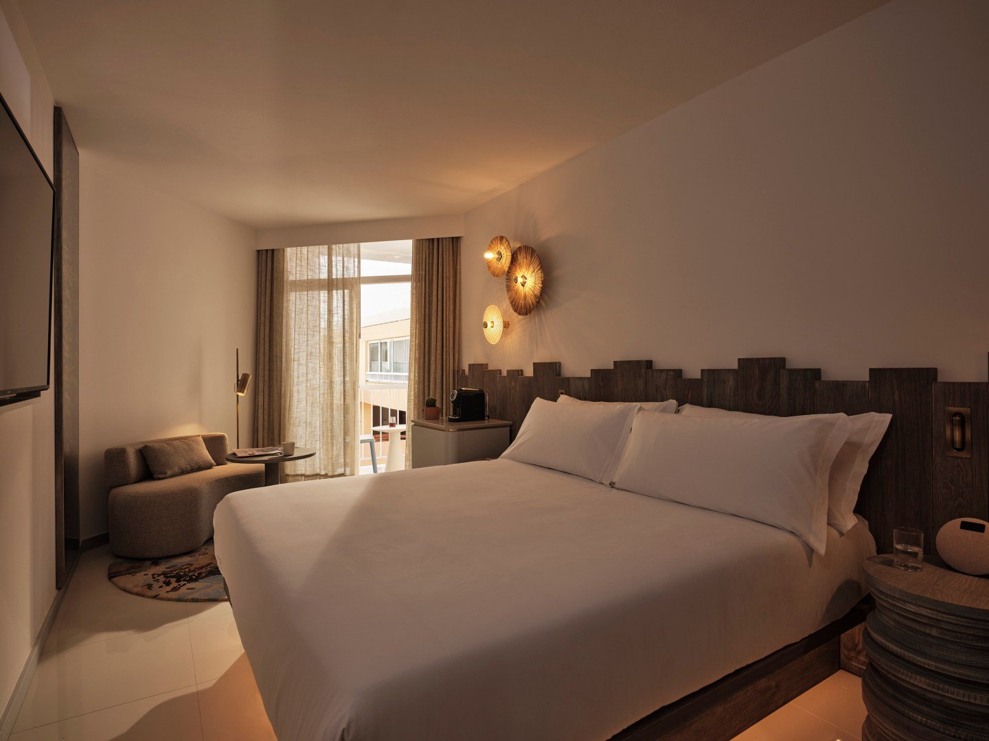 Modern hotel room with large bed, unique wooden headboard, soft lighting, couch, small table, and curtains with balcony view.