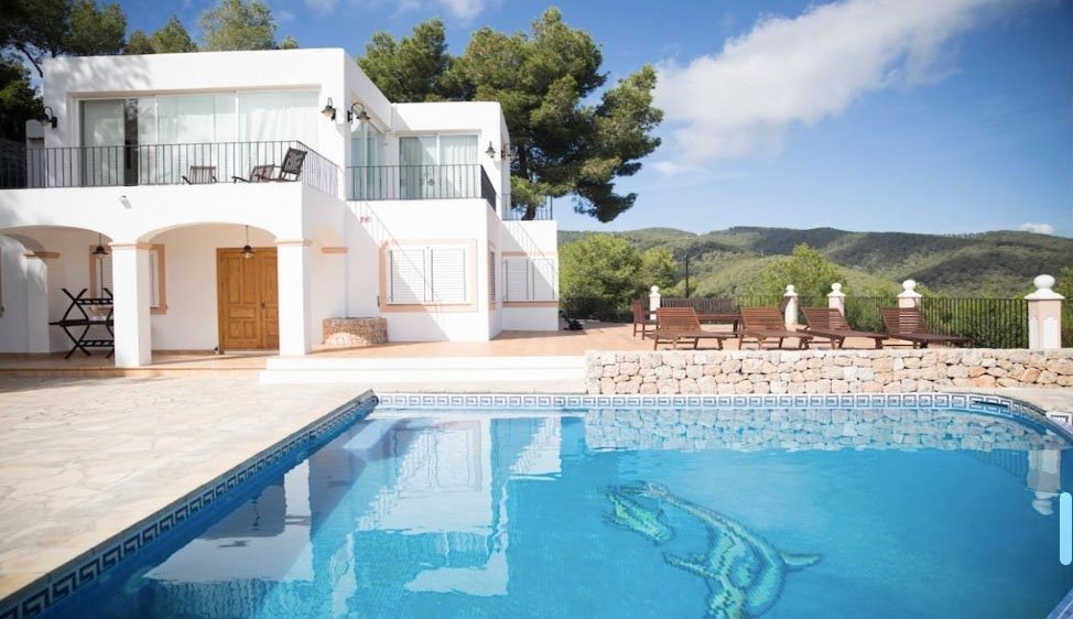 A modern white house with balconies and patio furniture, overlooking a swimming pool with a mosaic dolphin design, surrounded by mountains and greenery in the background.