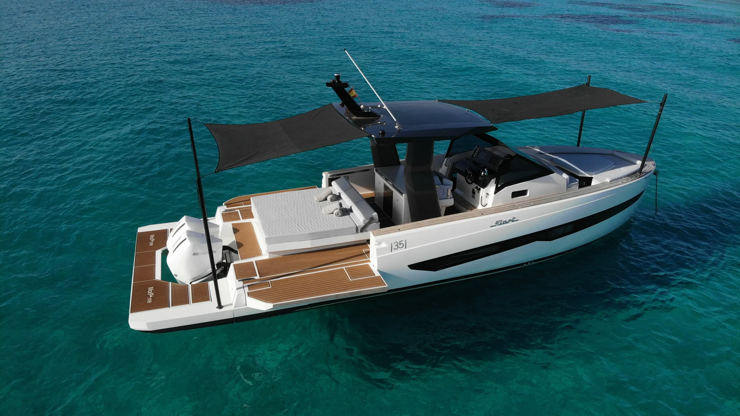 Aerial side view of the Fiart 35 Martone W with front and rear sunshades extended while anchored in shallow blue water.