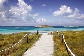 Wooden pathway leading to a beach with clear blue water and an island in the distance under a partly cloudy sky.