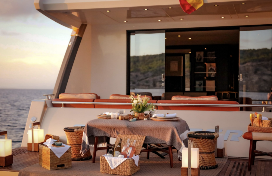 Luxury yacht deck with table set for dining, wicker baskets, and outdoor seating overlooking ocean.