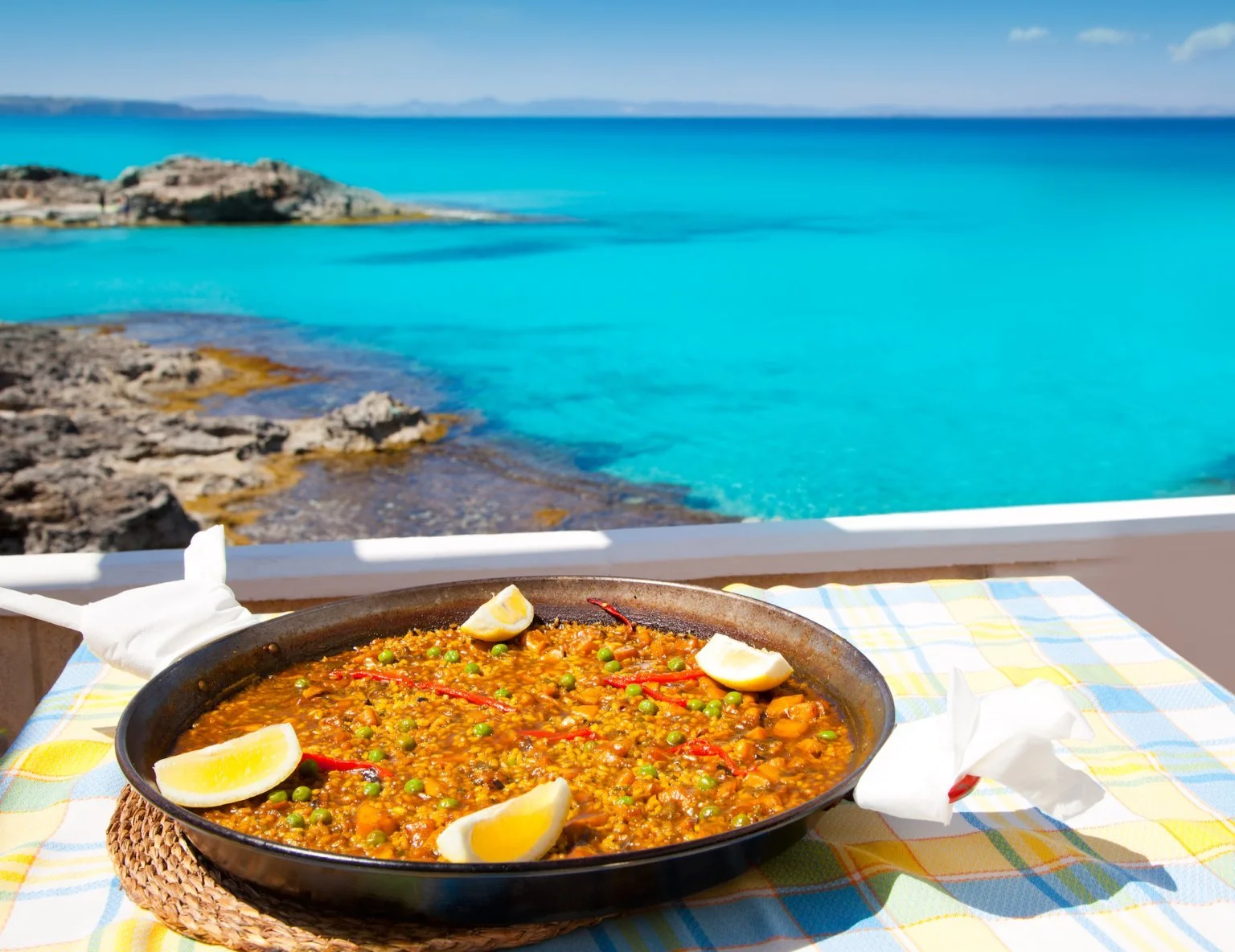 Pan of paella with lemon wedges on a table overlooking turquoise sea and rocky coastline.