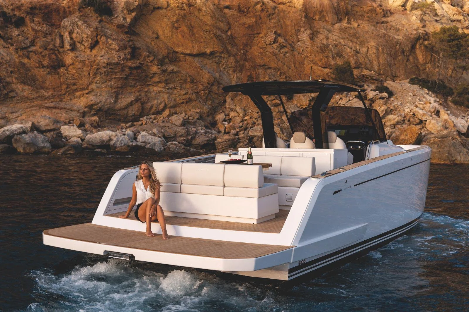 Luxury yacht on water with a person sitting on the deck, rocky coastline in the background.