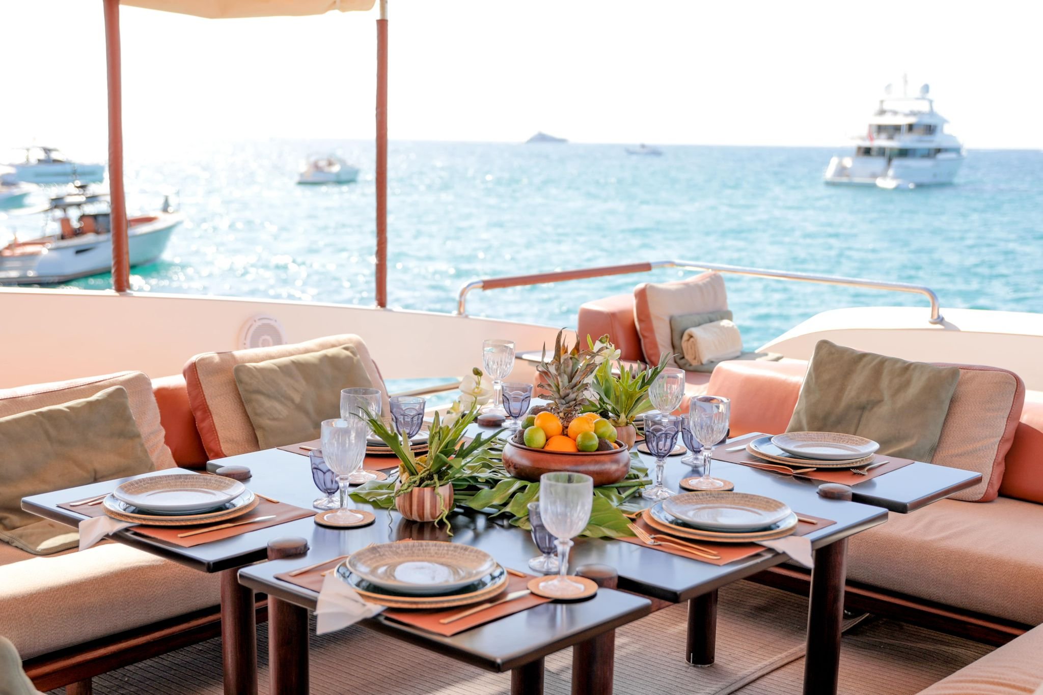 Luxury yacht deck dining setup with a table, plates, glasses, cushions, and ocean view with boats.