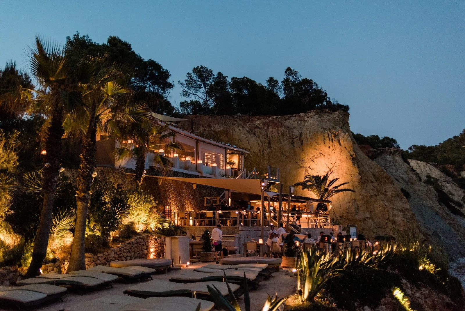 Outdoor restaurant with ambient lighting on a hillside with palm trees, surrounded by natural scenery.