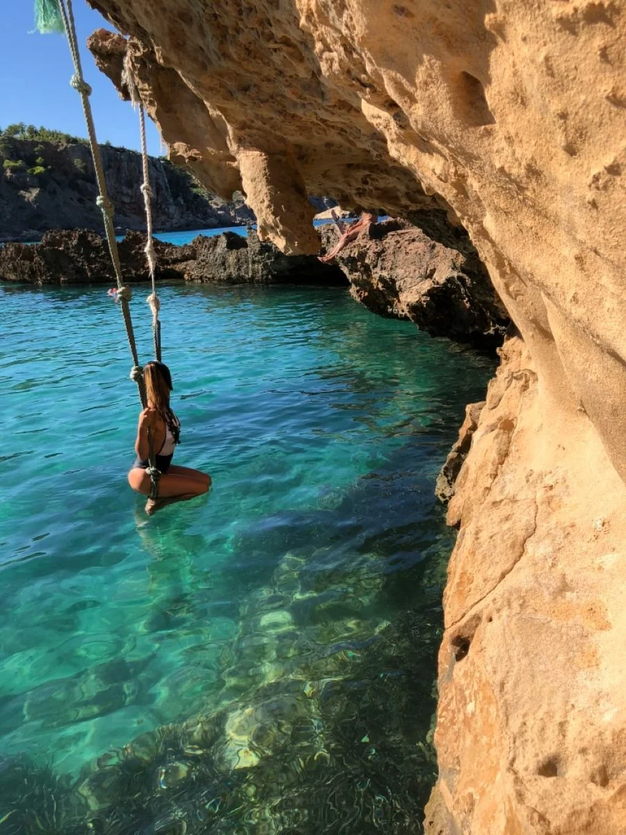 Person on swing over clear blue water near rocky coastline.