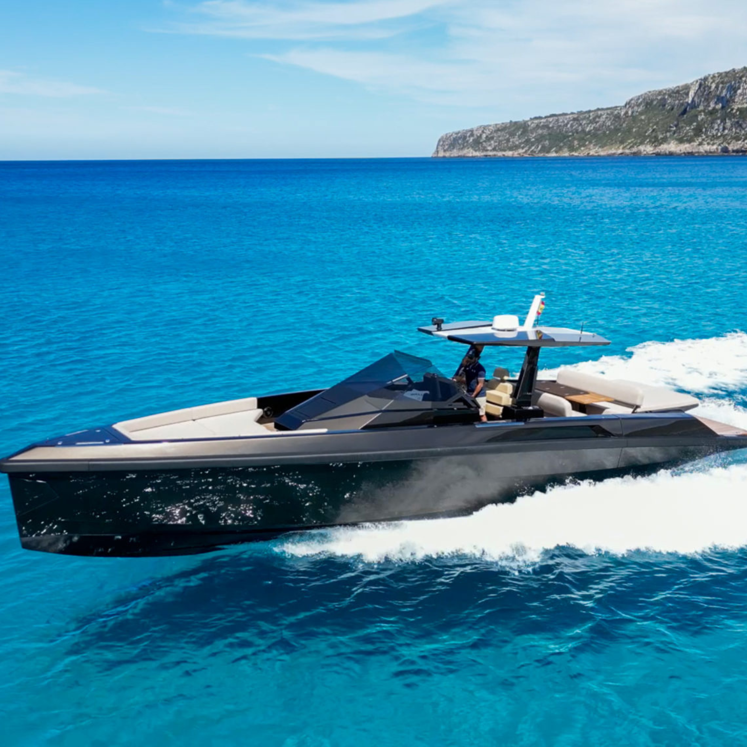 Sleek motor yacht cruising on clear blue water with rocky coastline in the background.