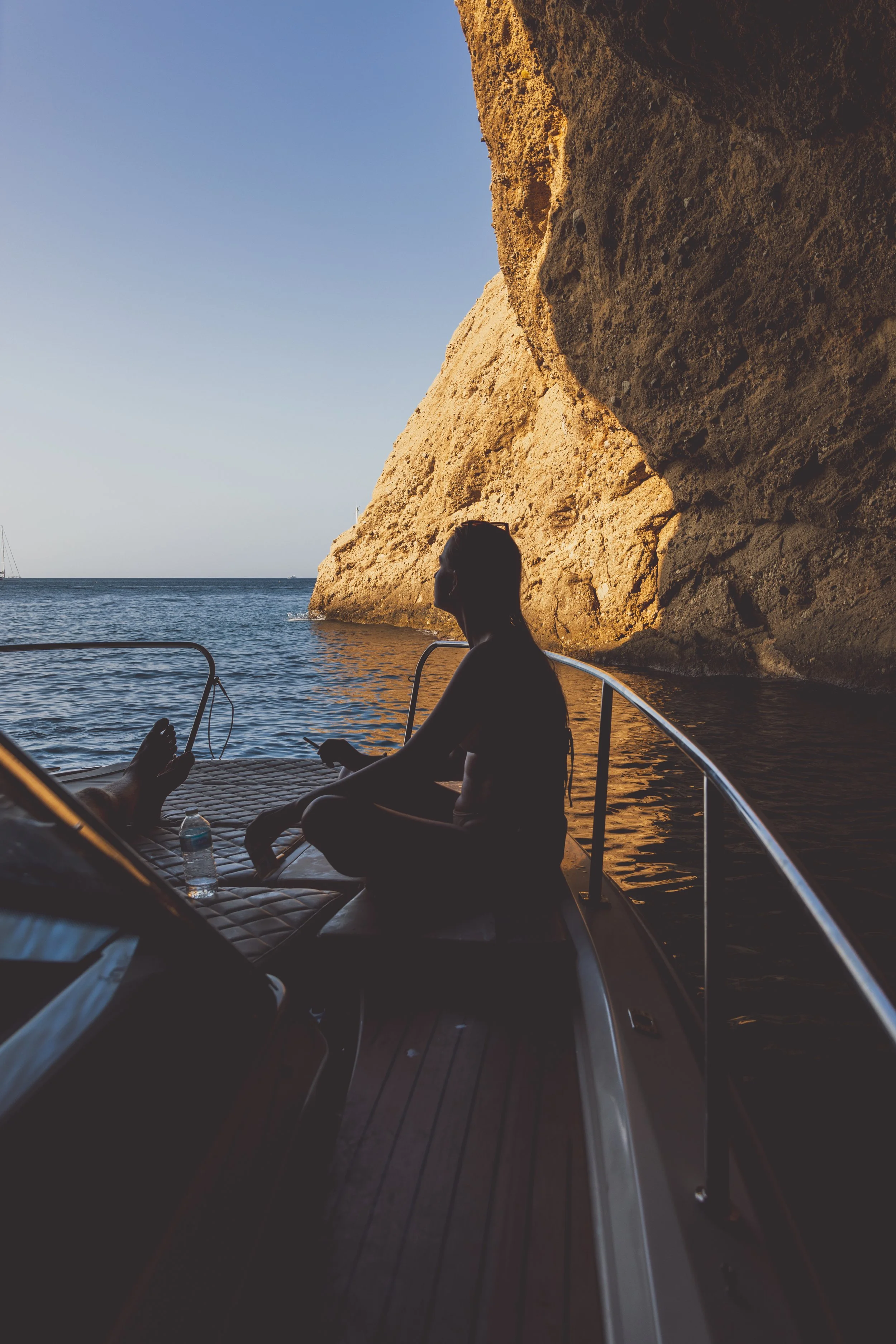 Person sitting on a boat near a rocky cliff in sunlight, overlooking the ocean.