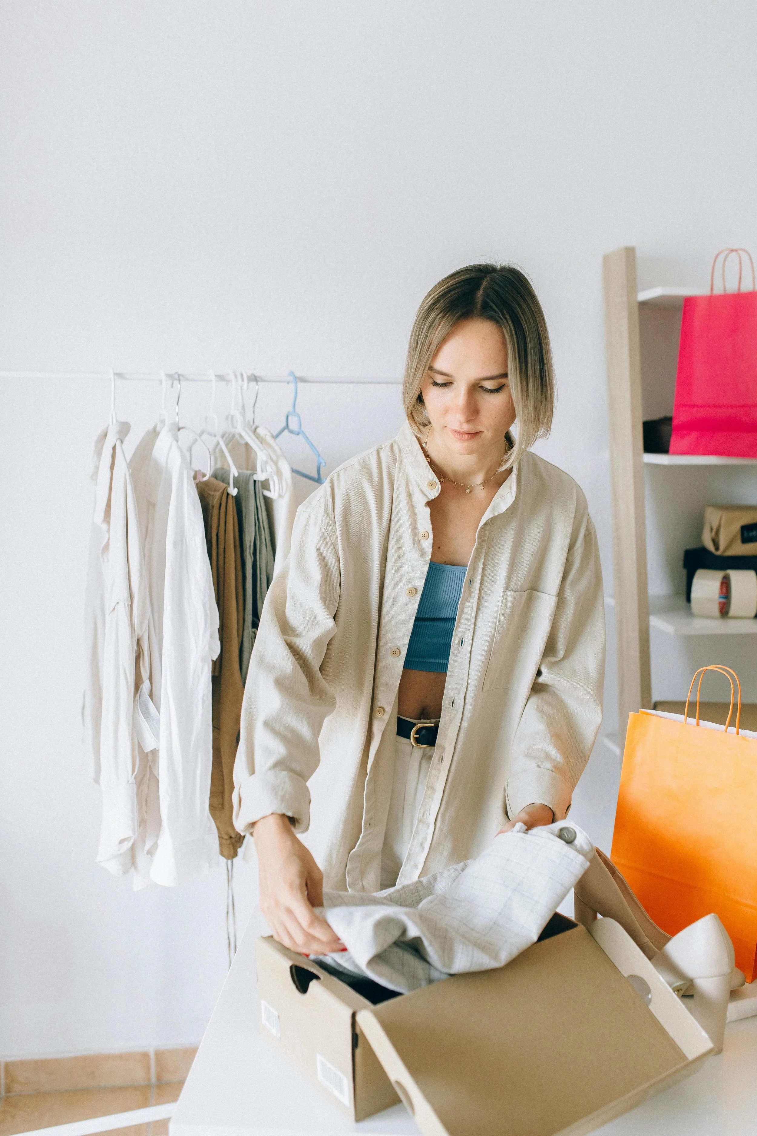 Woman in beige shirt unpacking clothes from a box, with clothing rack and shopping bags in the background.