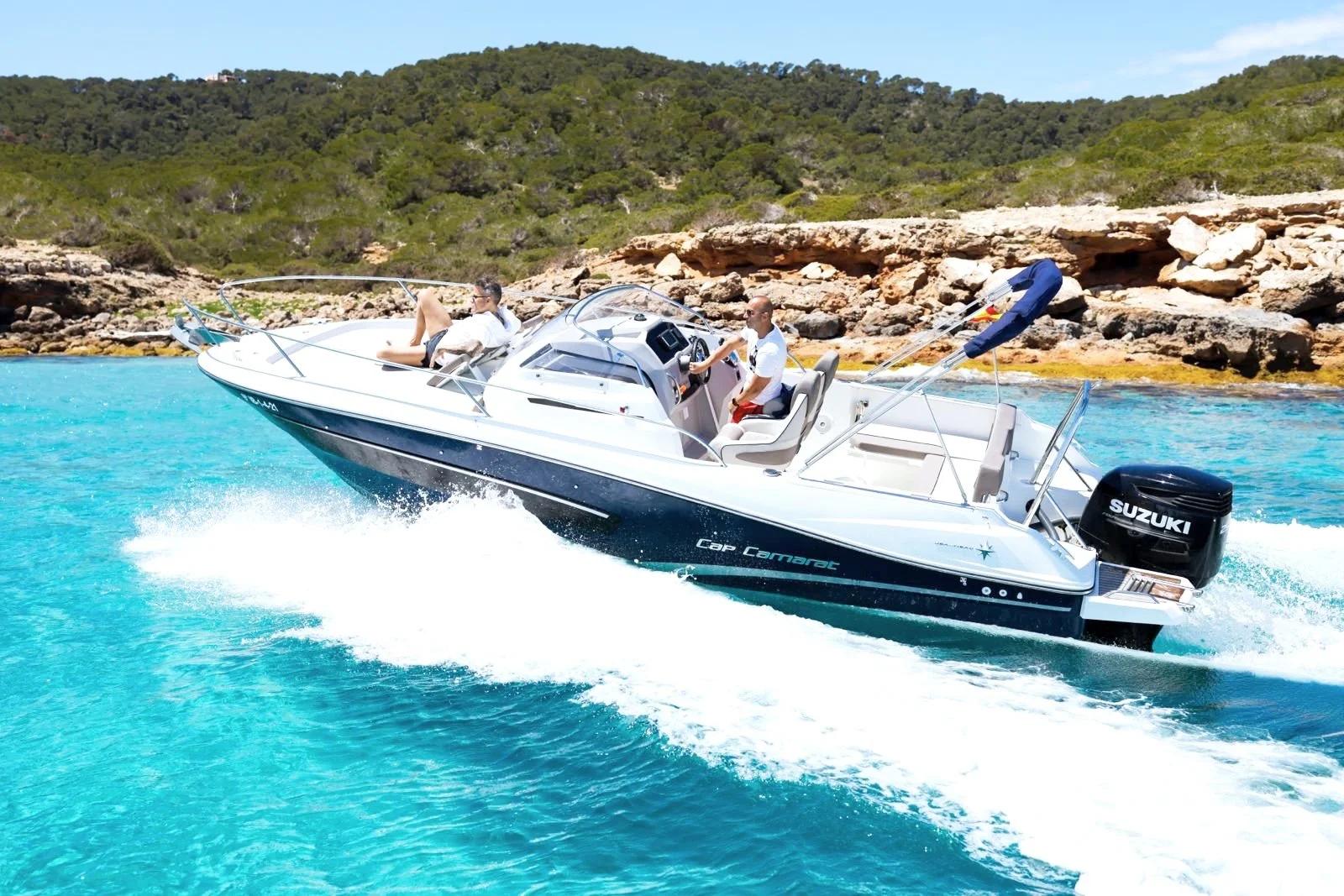 Motorboat with Suzuki engine in turquoise waters, two people on board, rocky shoreline and green hills in the background.