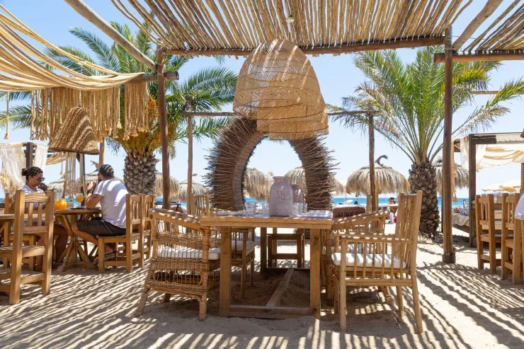 Beachside restaurant with wooden furniture and palm trees, decorated with straw canopies and natural materials.