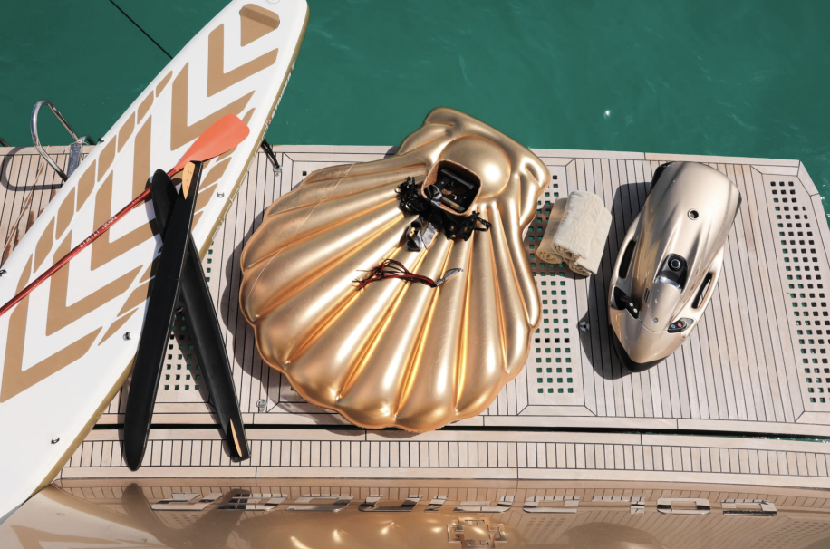 Stylish paddleboard with oars, a golden seashell-shaped inflatable, dive scooter on yacht deck near turquoise water.