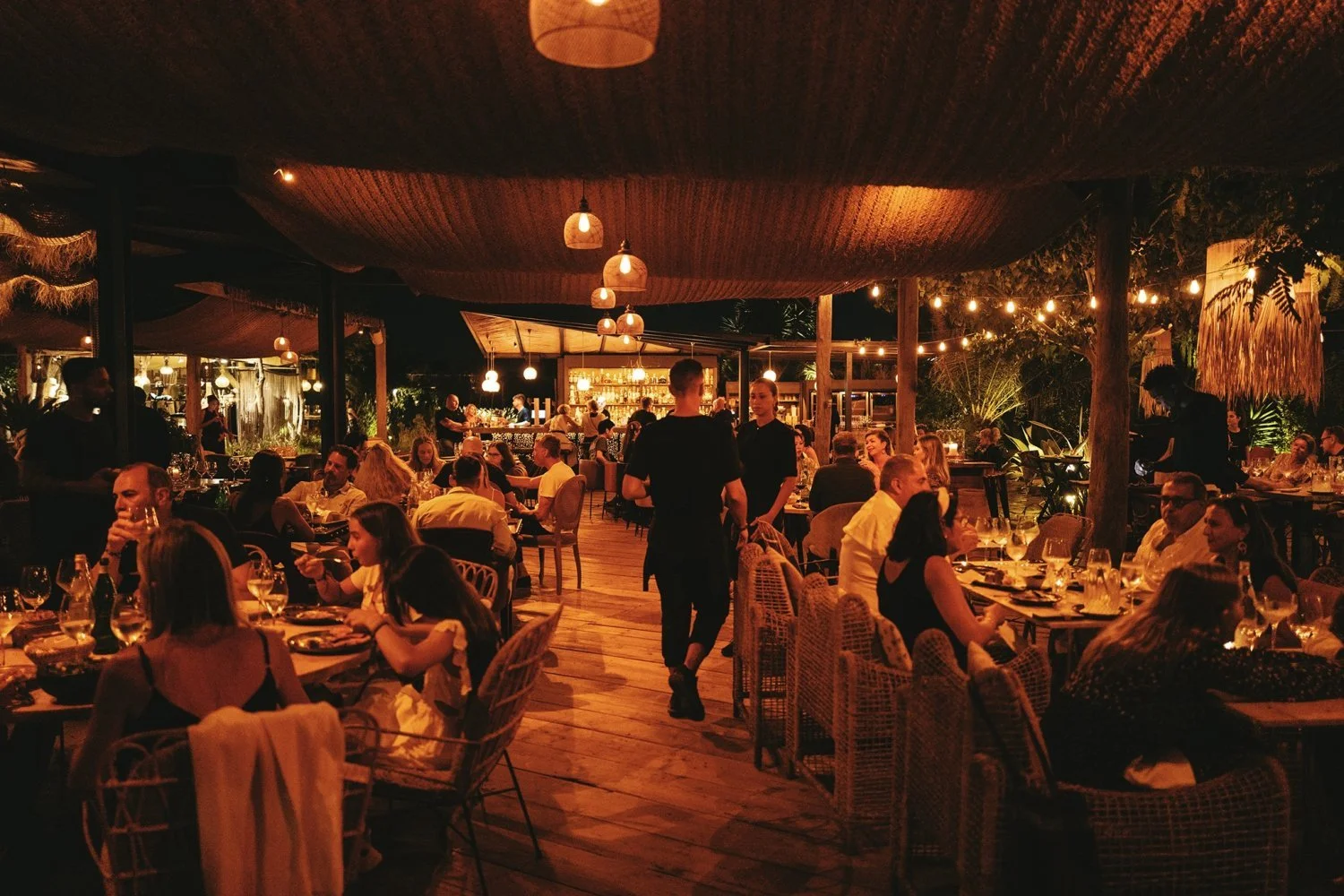 Outdoor restaurant with people dining at wooden tables, dimly lit with hanging lights and warm ambiance.