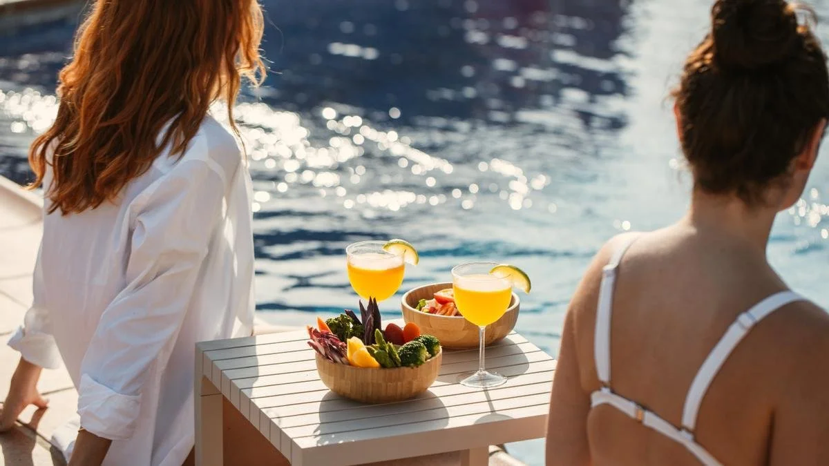 Two women by a swimming pool with fruit and drinks on a table.