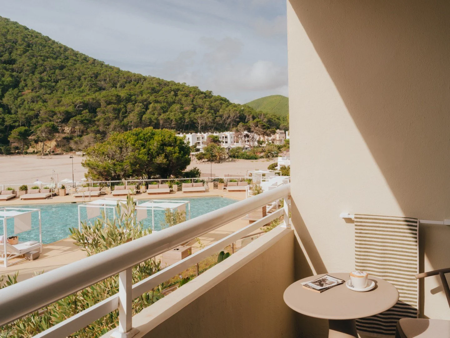 Hotel balcony with coffee cup on table, pool view, green hills, and resort buildings