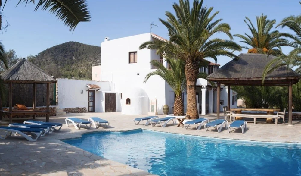 Resort with pool, palm trees, white buildings, and mountain in the background, sunny weather.
