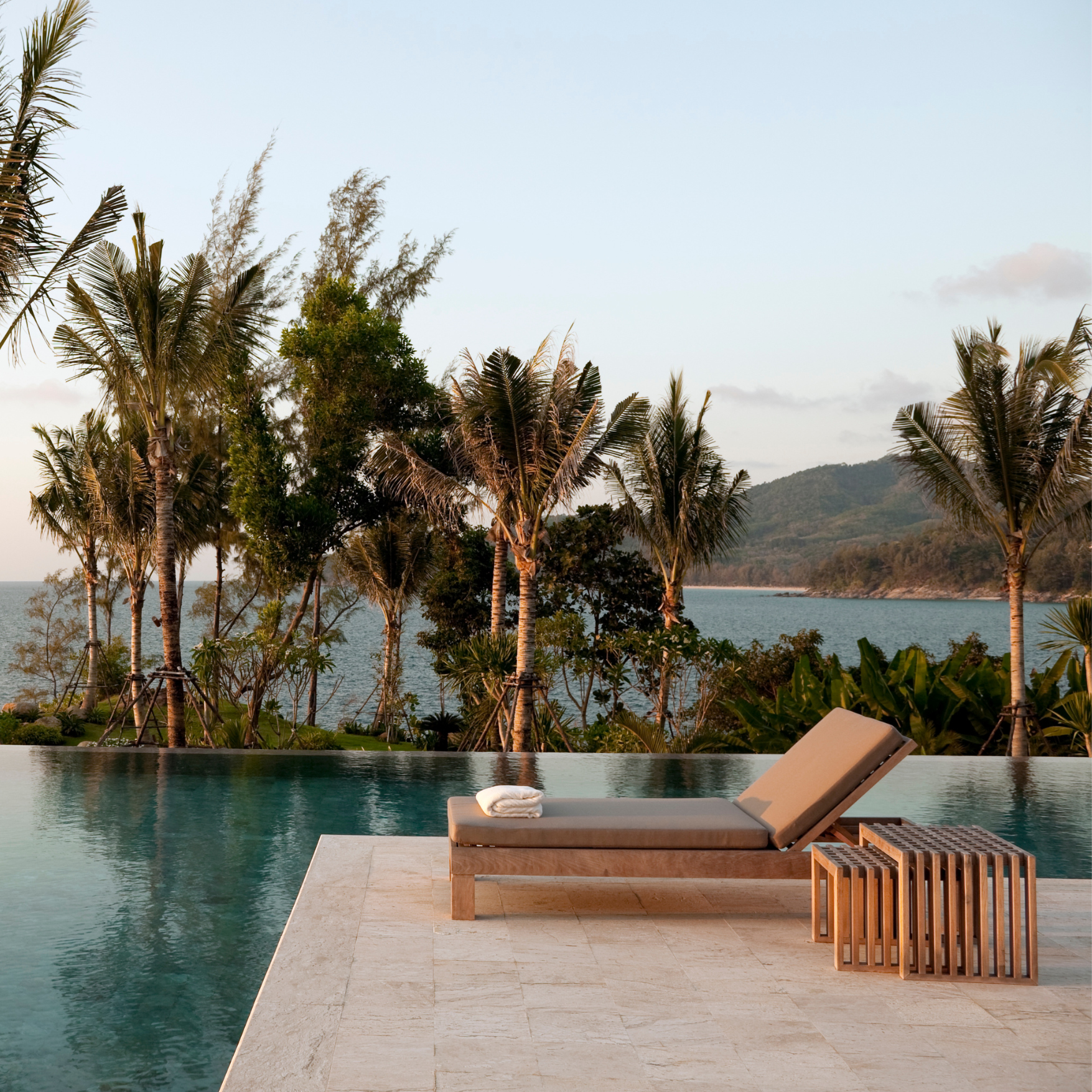 Lounge chair by an infinity pool with ocean and palm trees in background, tropical setting.