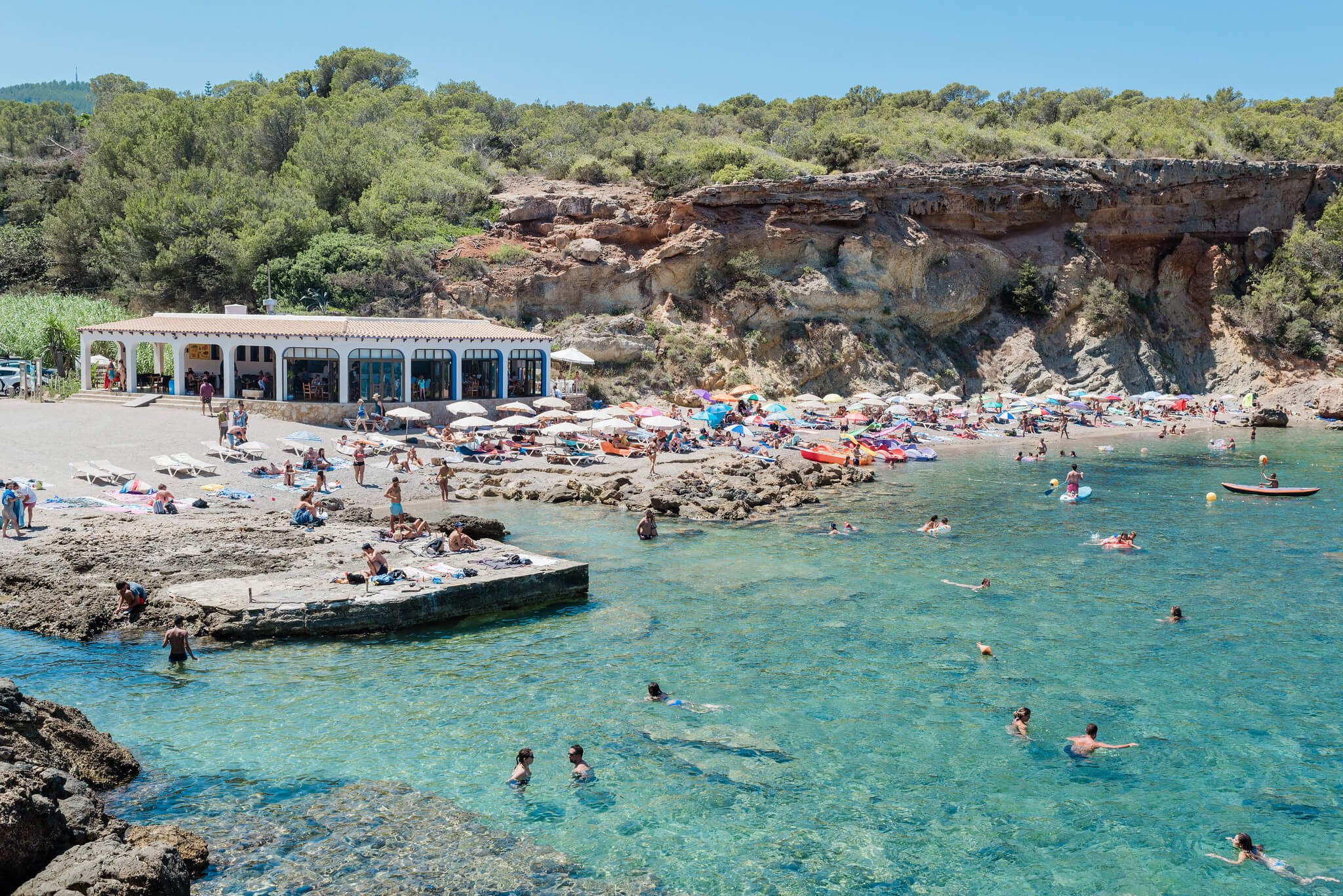 Beach scene with people swimming, sunbathing, umbrellas, and a white building; rocky shore and greenery in the background.