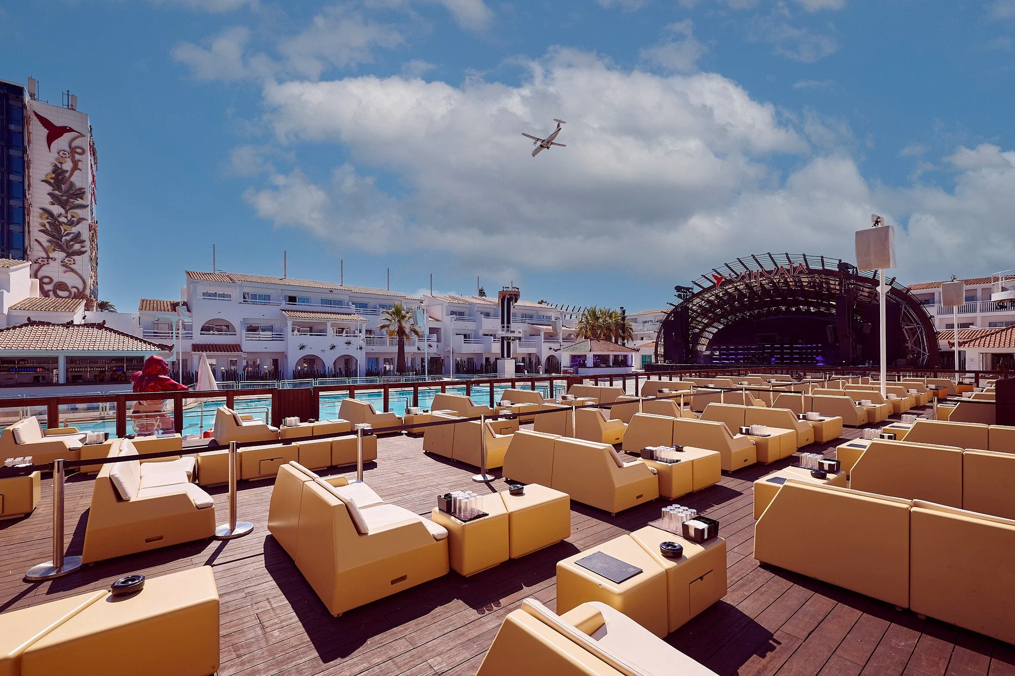 Outdoor lounge seating facing a stage, with a swimming pool and buildings in the background, under a blue sky with an airplane overhead.