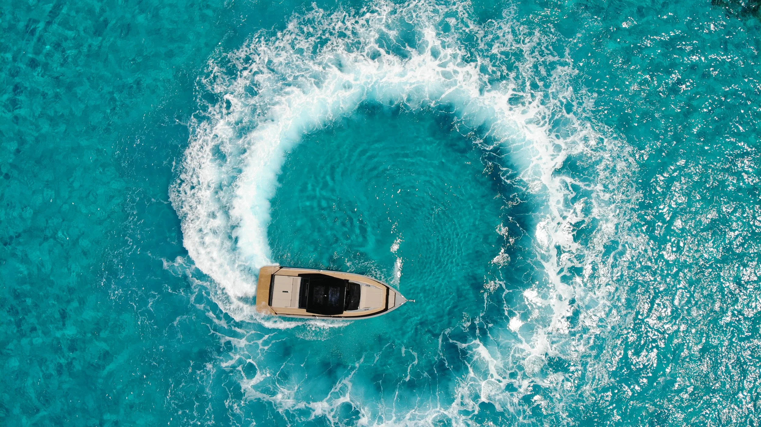 A boat creating a large circular wake pattern in bright blue ocean water seen from above.