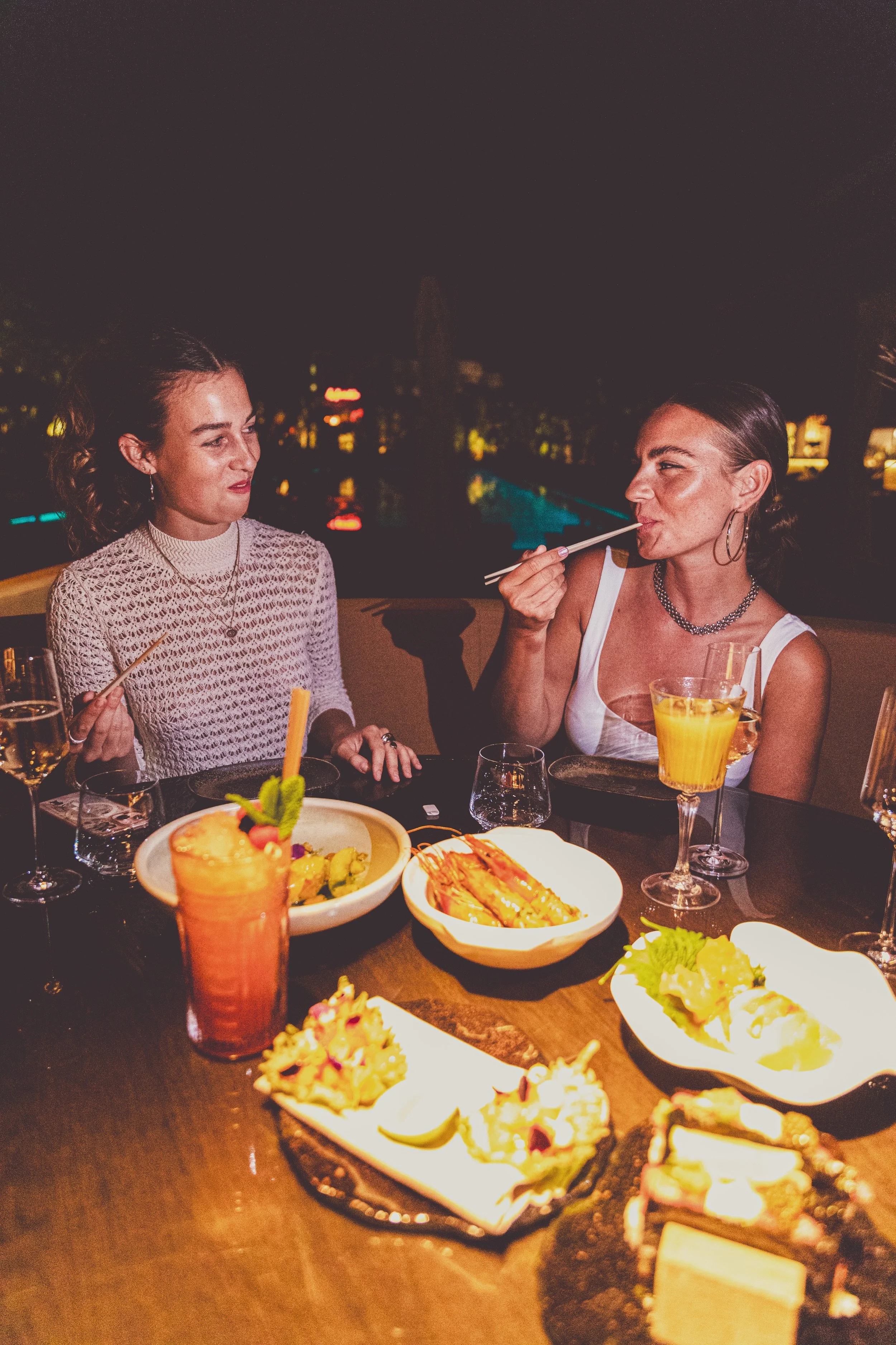 Two women dining at a restaurant table with various dishes and drinks at night.