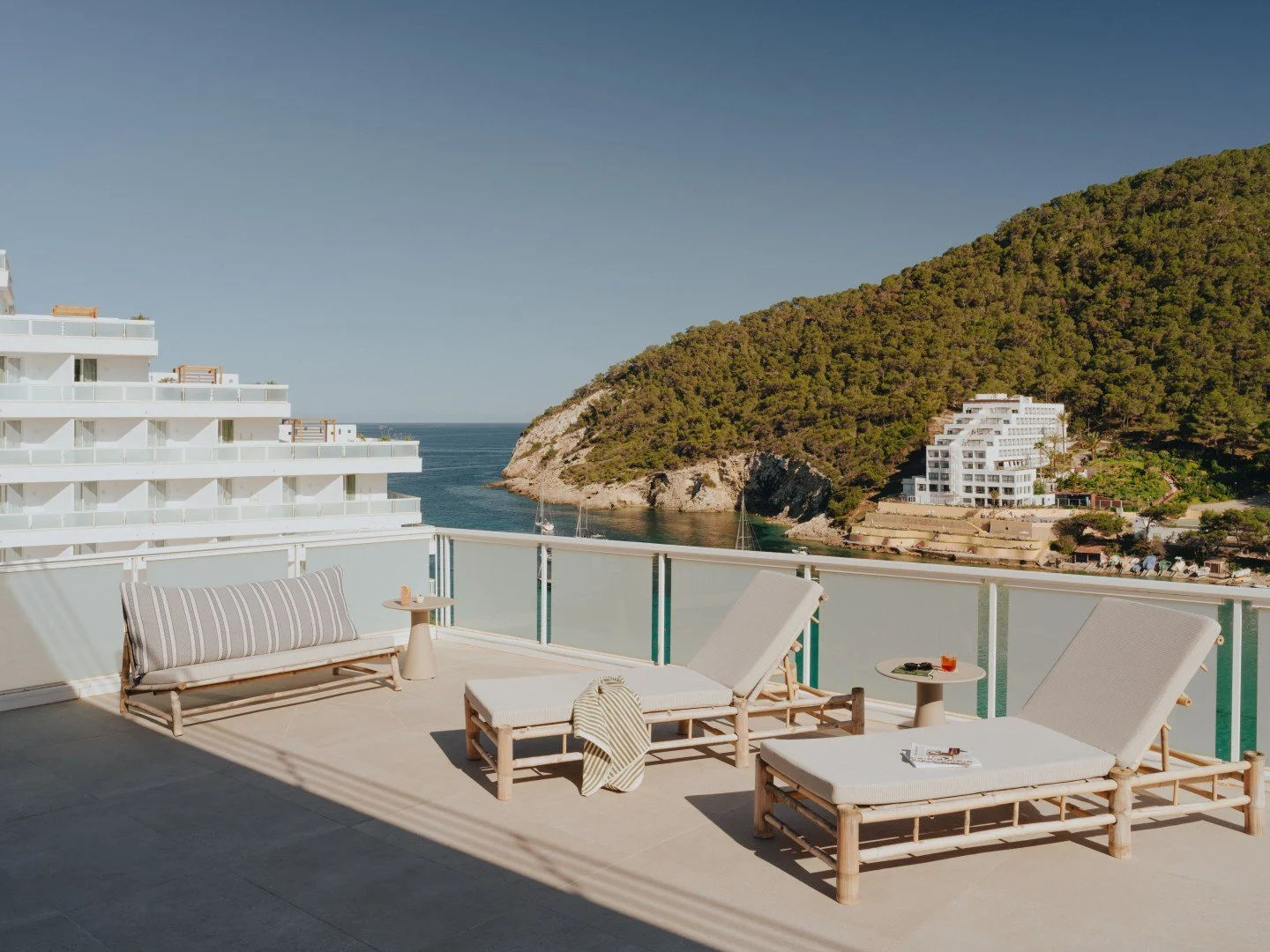 Outdoor terrace with lounge chairs, table, and sea view with hillside and white buildings.