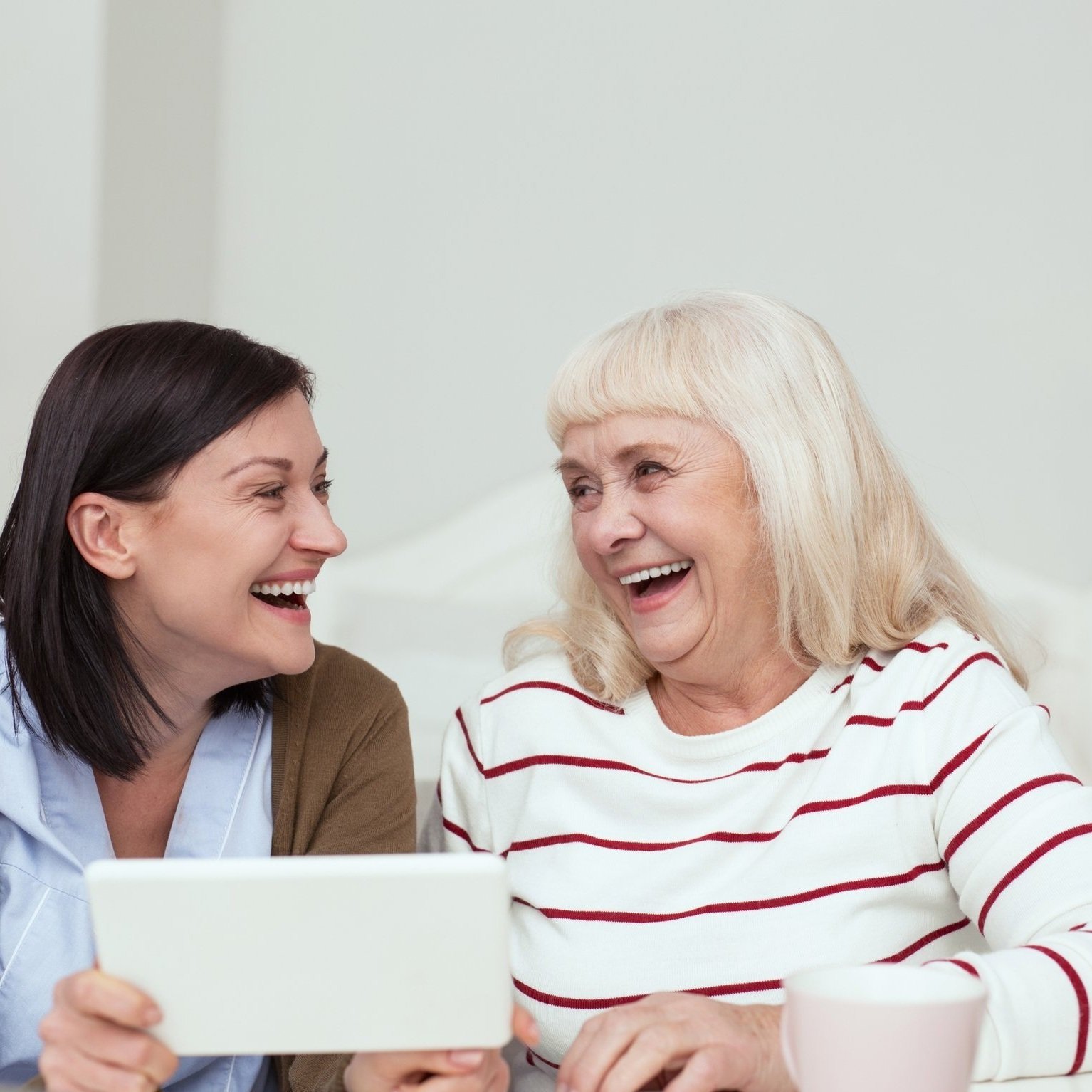 Eine junge braunhaarige Frau und eine ältere blonde Frau lächeln sich an. und sitzen vor einem weißen Tablet.