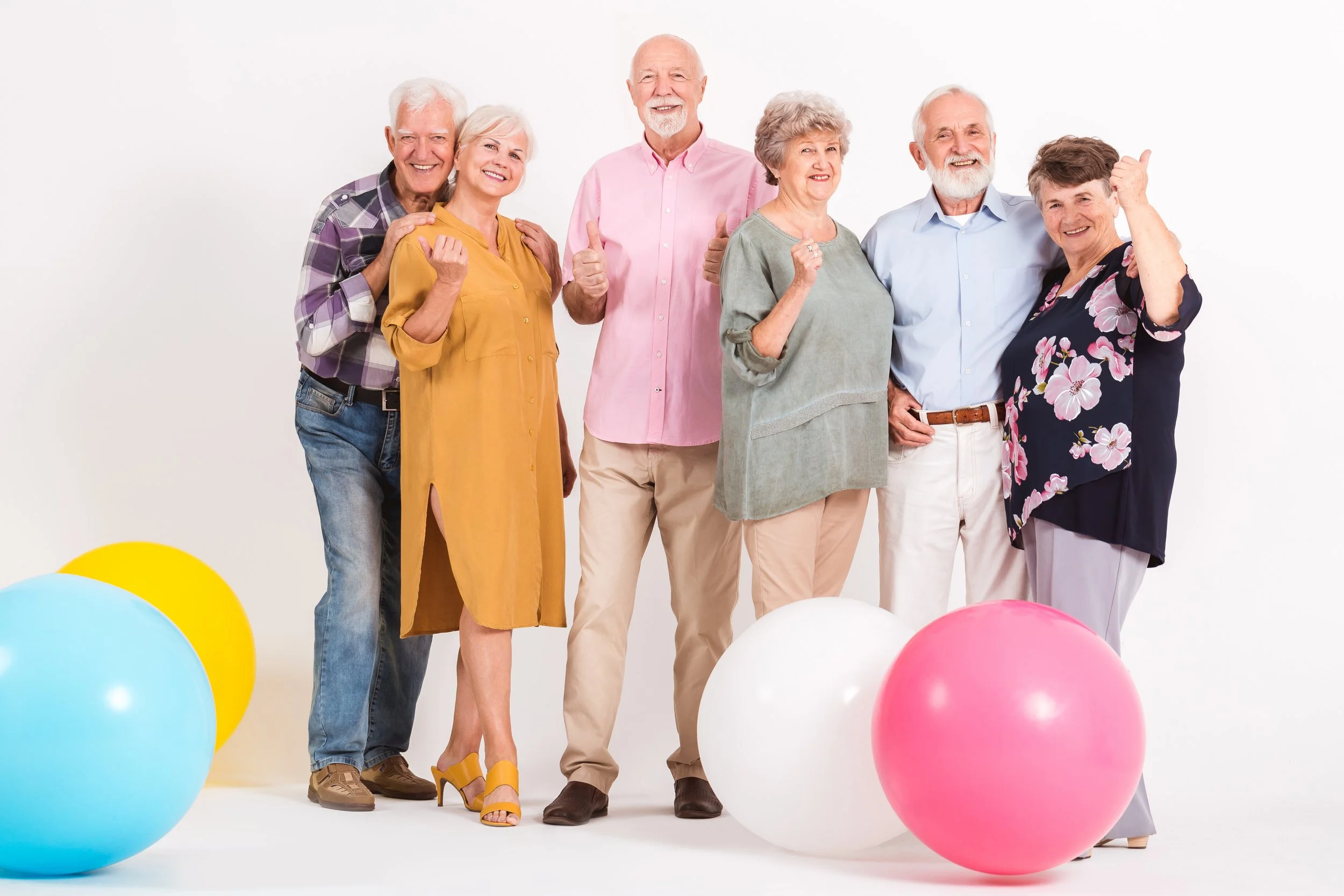 Drei Senioren und drei Seniorinnen stehen vor der Kamera, lächeln. Drei zeigen mit dem Daumen nach oben. Rechts im Bild sind Luftballons in Pastellfarben.