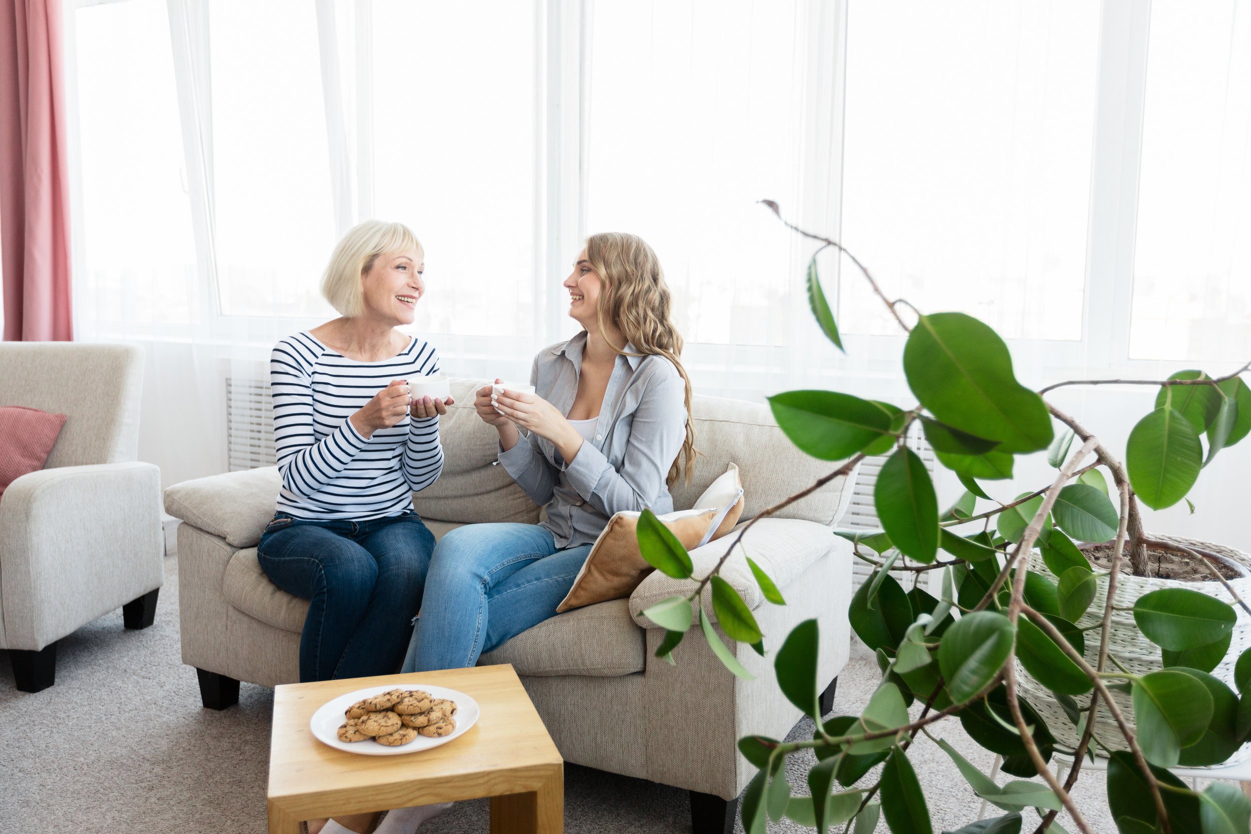 Zwei Frauen sitzen auf einem Sofa, lachen und trinken Kaffee, im Vordergrund eine große Pflanze, auf einem kleinen Tisch Kekse.