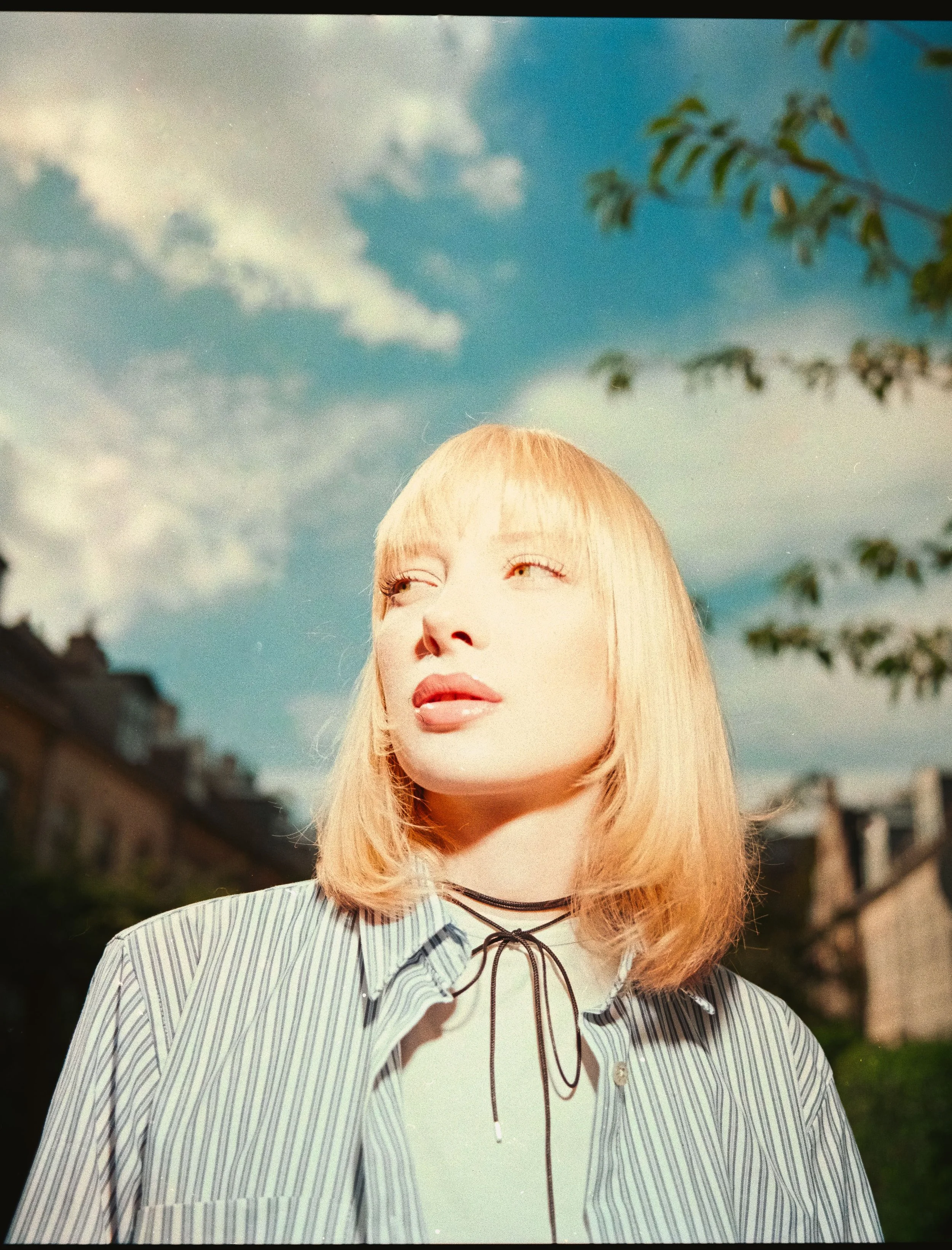 Young woman with blonde hair and a striped shirt standing outdoors on a sunny day, with blue sky and white clouds in the background.