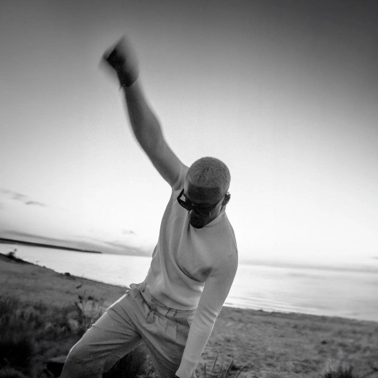 A black and white photo of a person wearing sunglasses and a long-sleeved shirt, bent forward with one arm raised and fist clenched, standing outdoors near water.