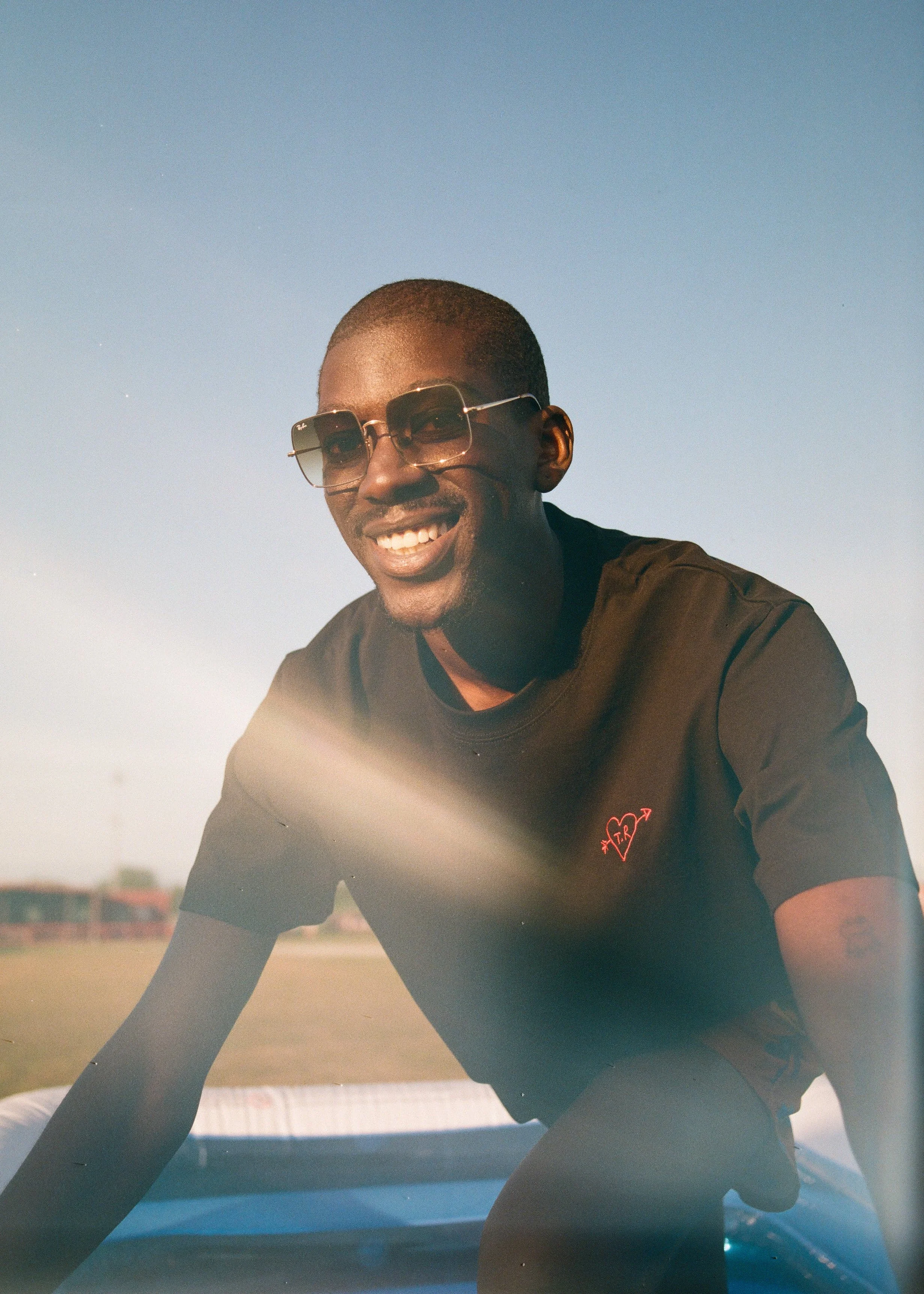 A smiling man wearing sunglasses and a black T-shirt with a red logo, leaning forward outdoors during daytime with a clear blue sky in the background.