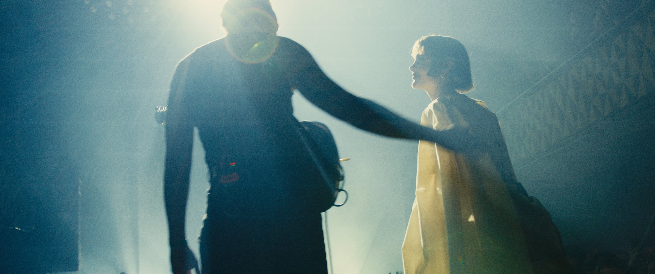 Two people, a man and a woman, holding hands in a spotlighted, hazy setting, likely a stage or concert.