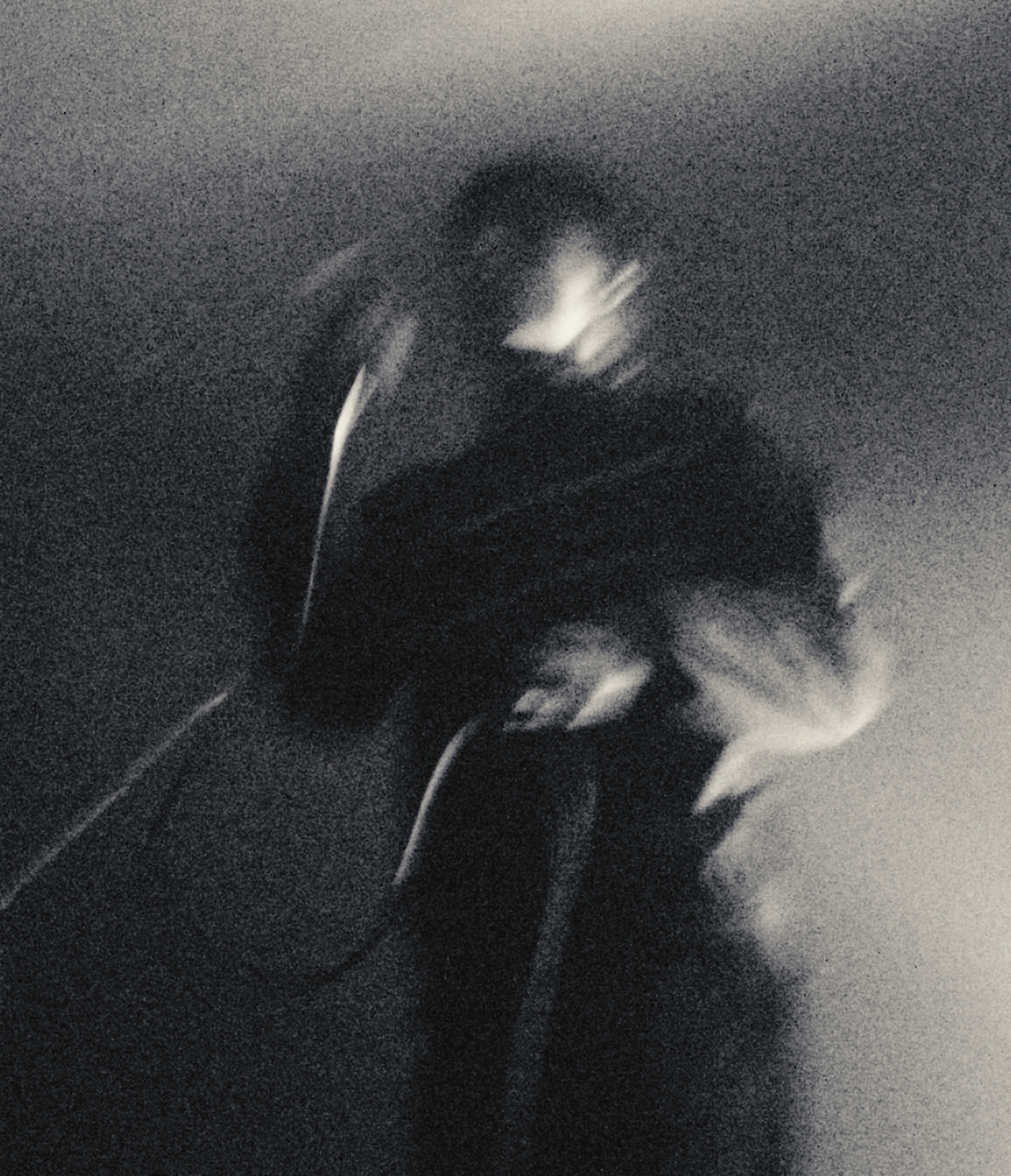 A black and white blurred self-portrait of a person holding a camera or phone to take a picture, with a hand near their face and long, dark hair, against a plain wall.