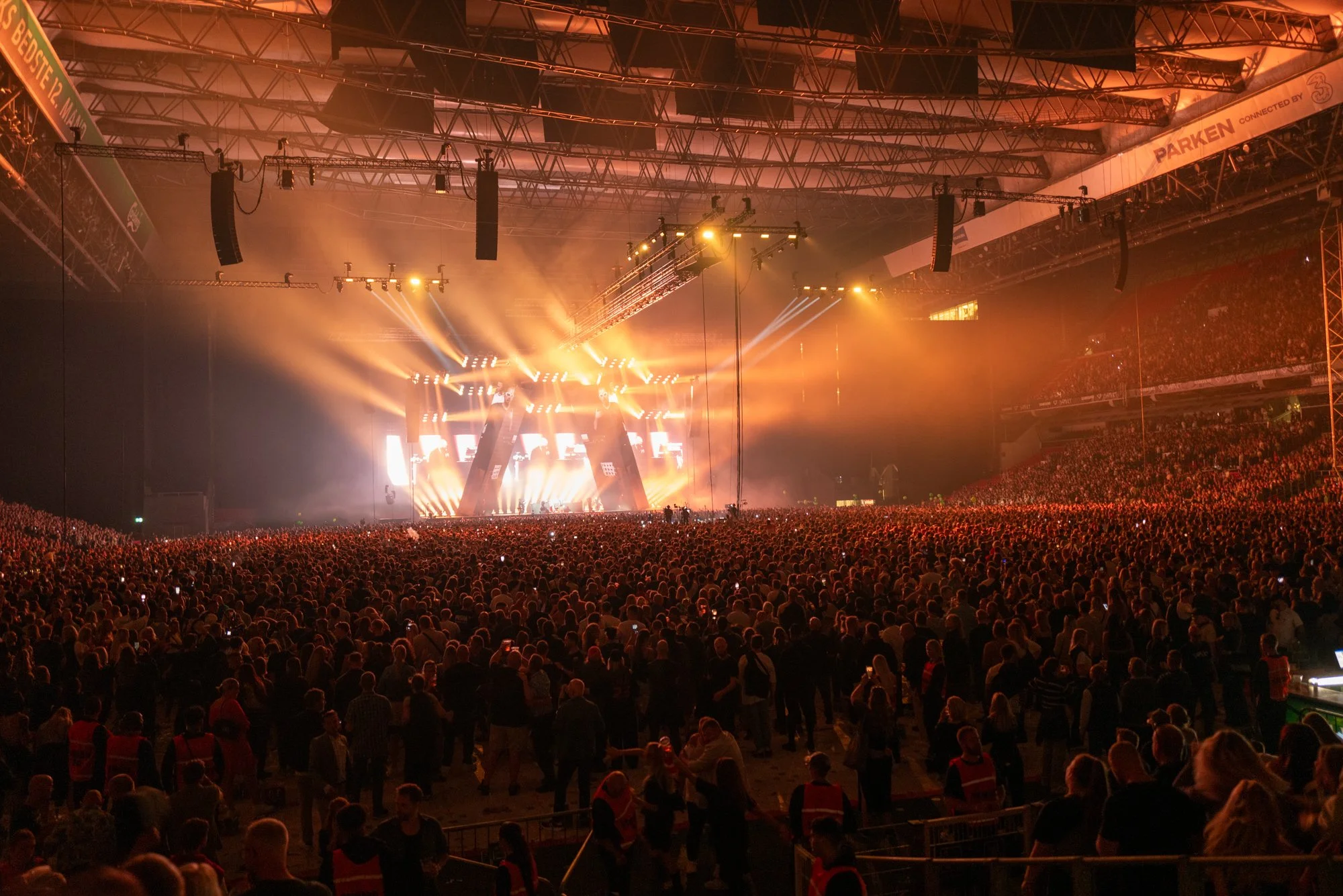 A large indoor concert or music festival with a massive crowd facing a brightly lit stage, with orange and yellow stage lights and screens at the back.