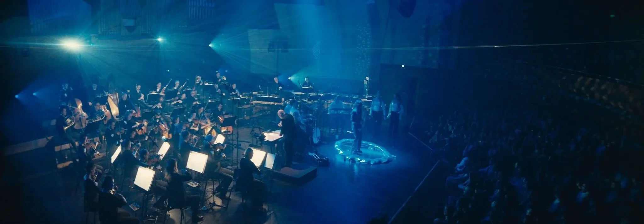 An orchestra performing on stage with blue lighting and a large audience in the background.