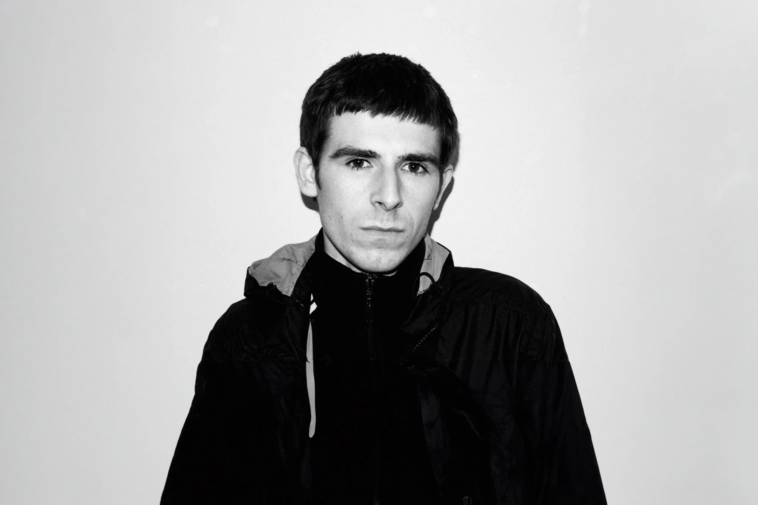 Black-and-white portrait of a young man with short dark hair, wearing a zip-up jacket, looking directly at the camera with a serious expression.