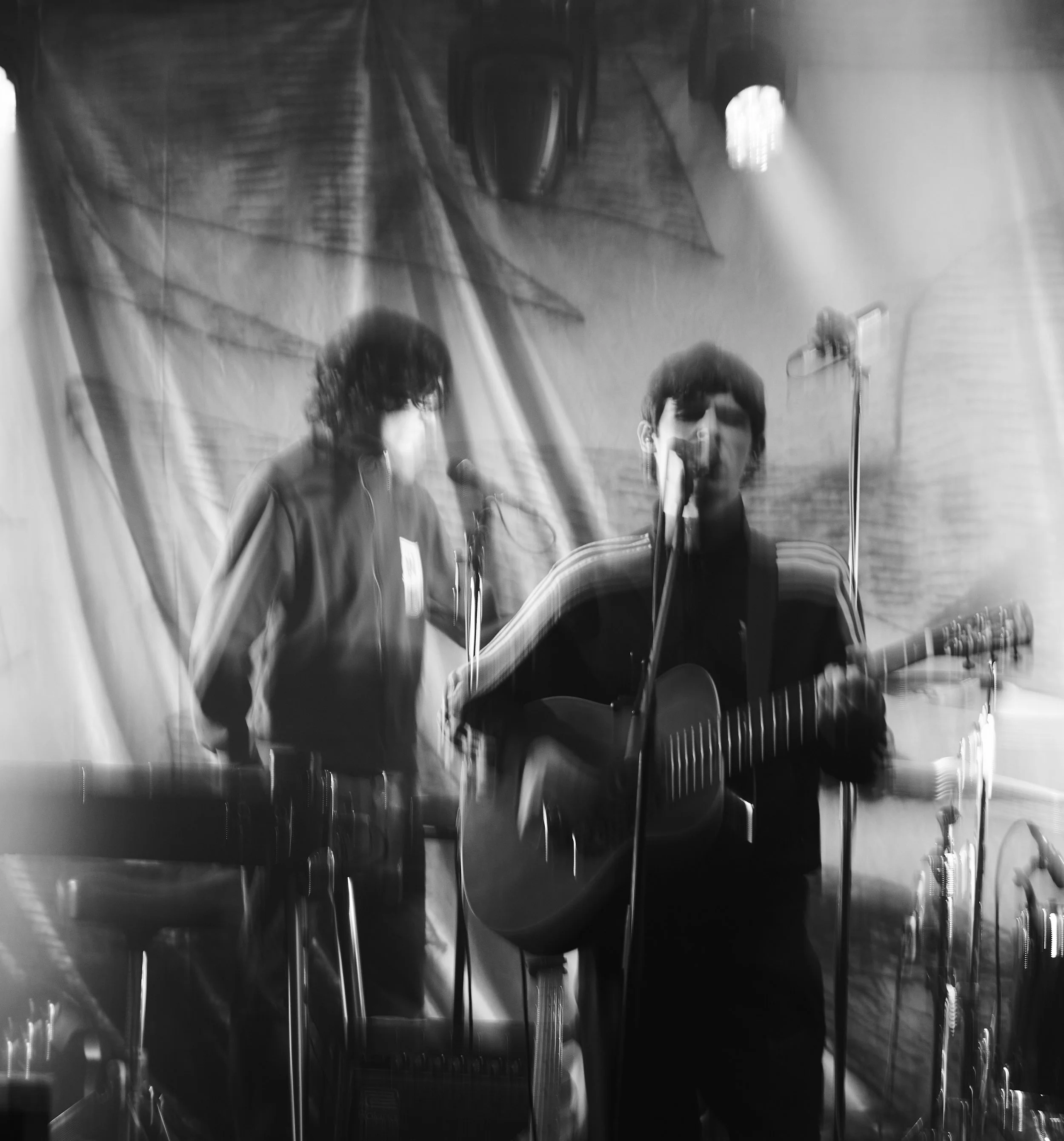 Two musicians perform on stage; one plays guitar and sings into a microphone, the other stands nearby with a keyboard; stage lighting creates a blurry, artistic effect.