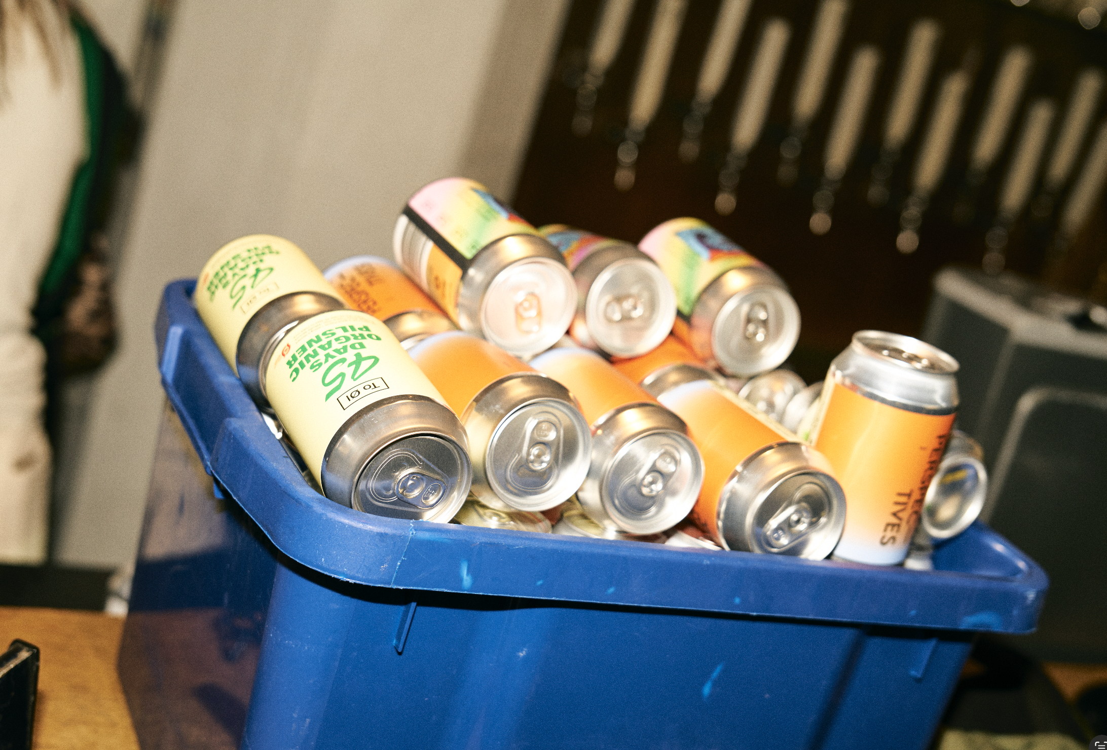 A blue plastic bin filled with cans of sparkling beverage, with some cans having a yellow and orange label. The cans are mostly aluminum and are piled inside the bin.