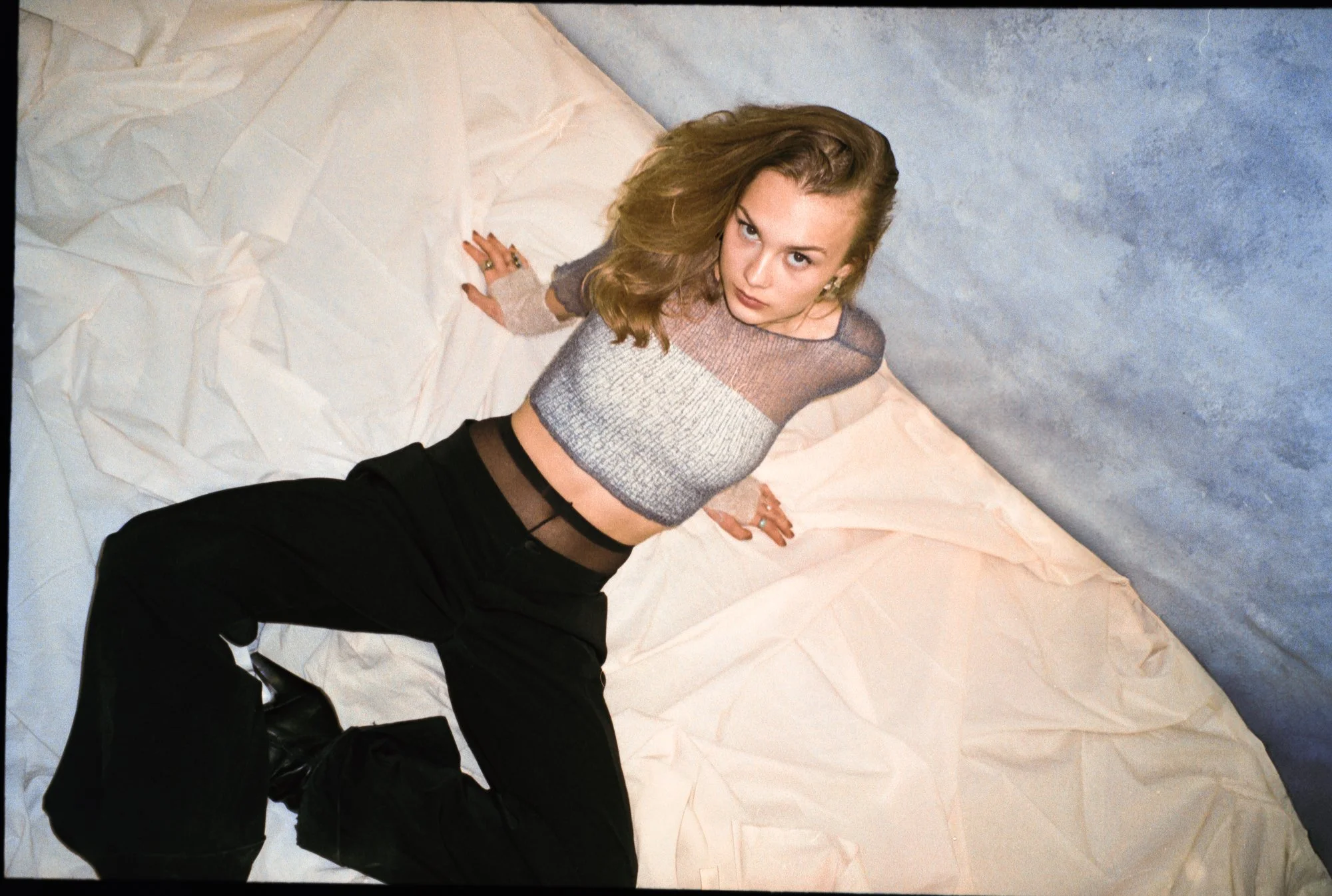 A young woman with wavy light brown hair looking up at the camera, posing on crumpled white and beige paper with a textured blue-gray background.