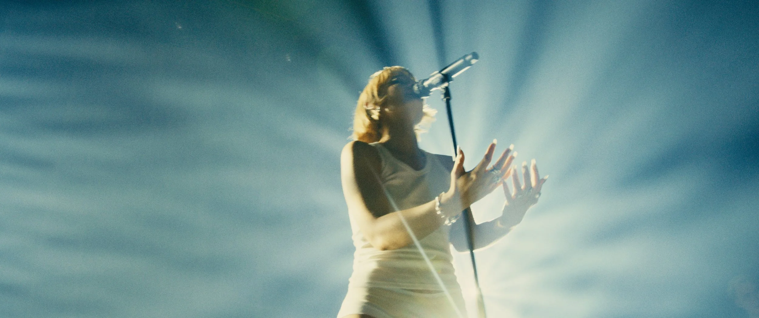 A person singing into a microphone on stage, with bright lights creating a dramatic effect.